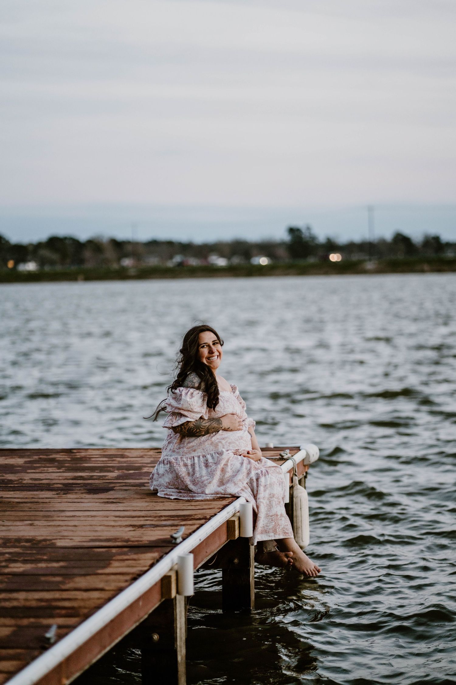 A person in a white dress sits on a wooden dock overlooking a serene lake at dusk.