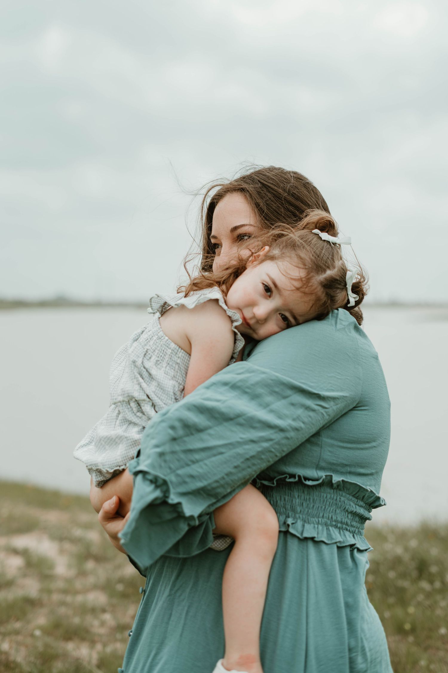 Two people share a tender embrace in matching teal outfits against a cloudy waterfront landscape.