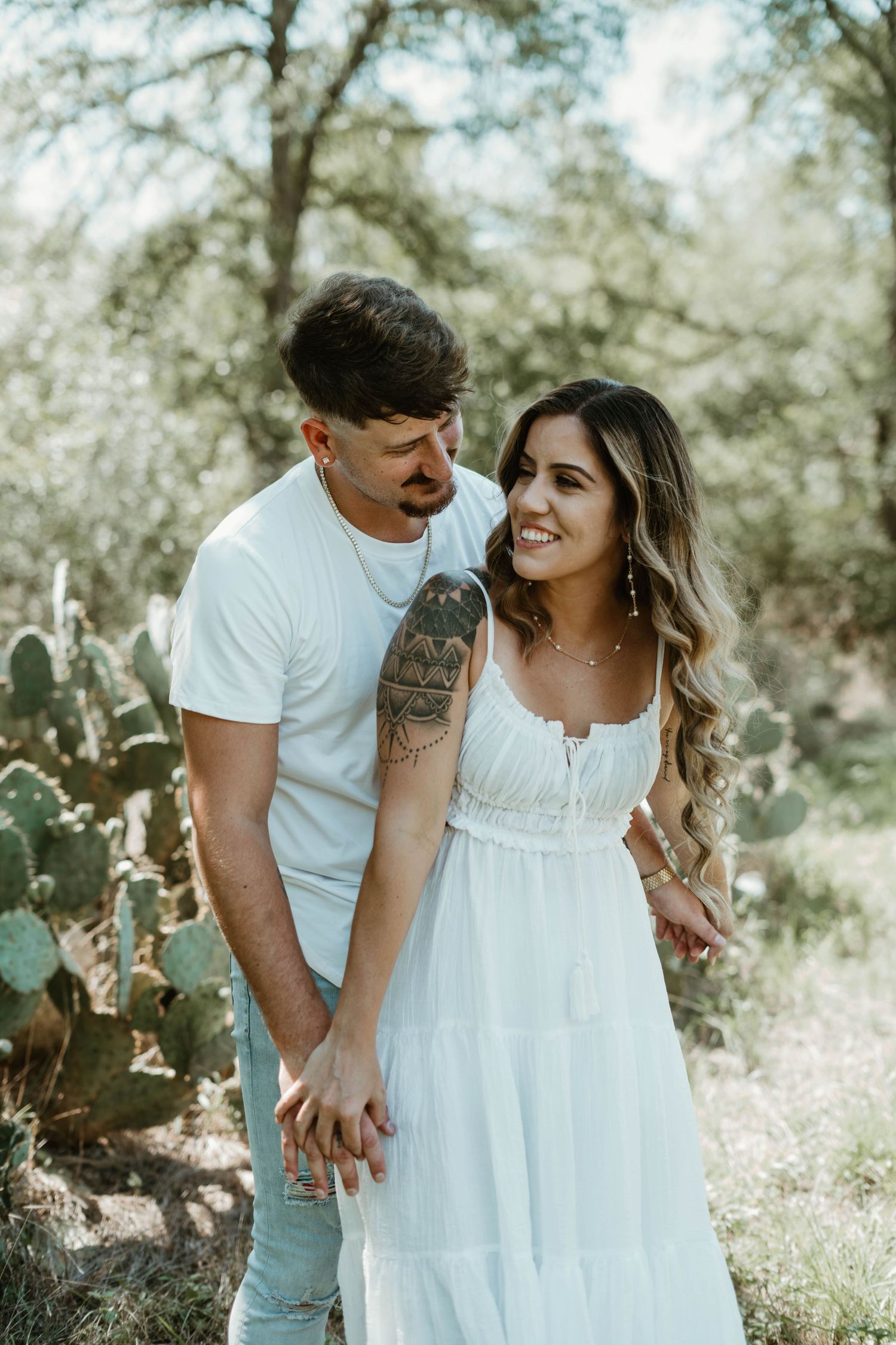 Couple sharing intimate moment in white outfits among cactus plants and trees in dreamy natural outdoor setting.