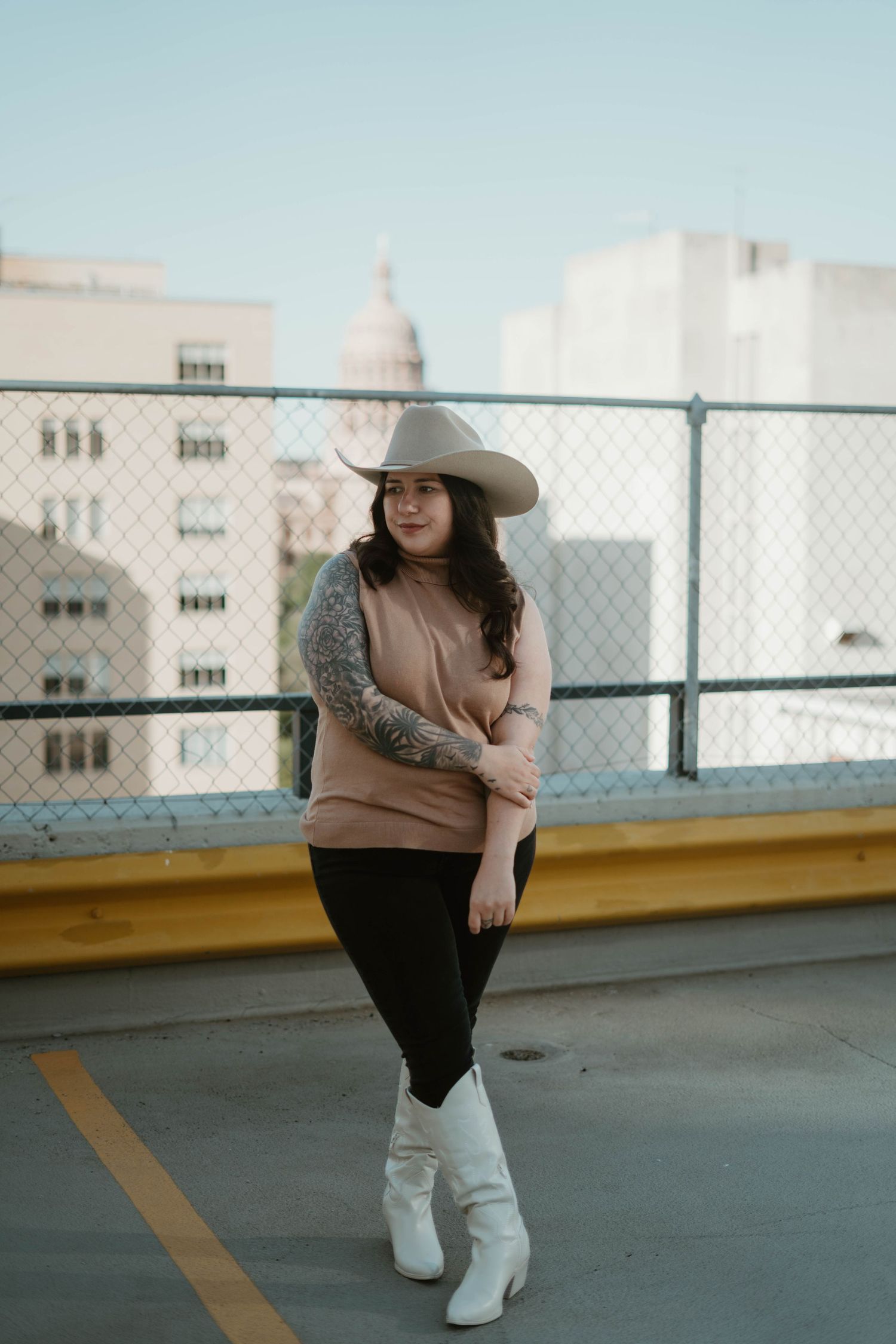 A person in a wide-brimmed hat, beige sweater and white boots poses on a rooftop parking area against a city backdrop.