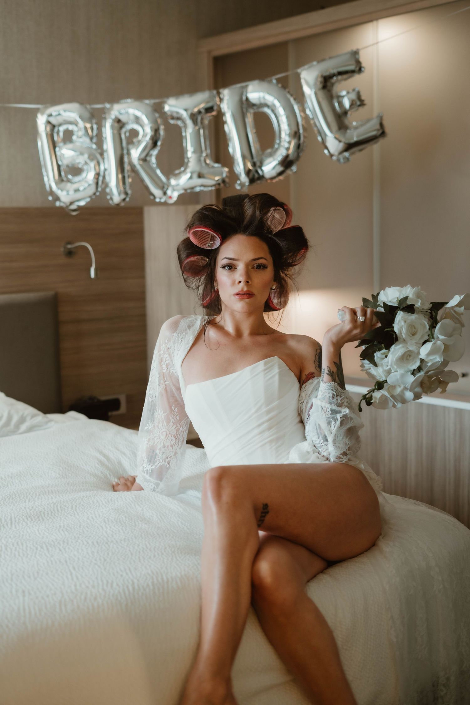 A bridal boudoir photo featuring hair rollers and a white dress against silver BRIDE balloon letters in the background.