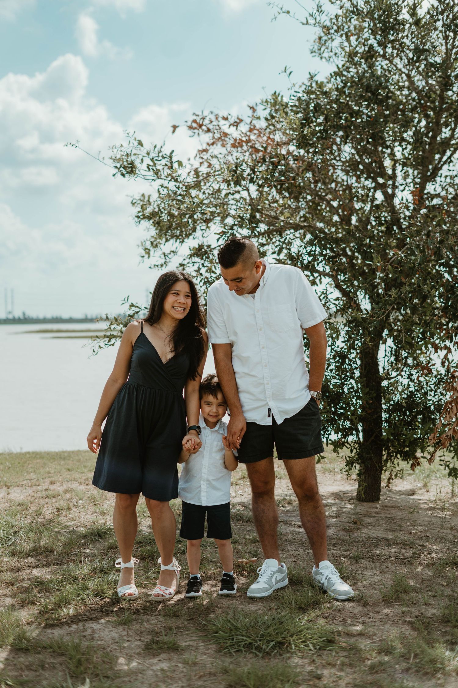 A family enjoys a sunny day together by a lake, standing under a leafy tree on the shoreline.