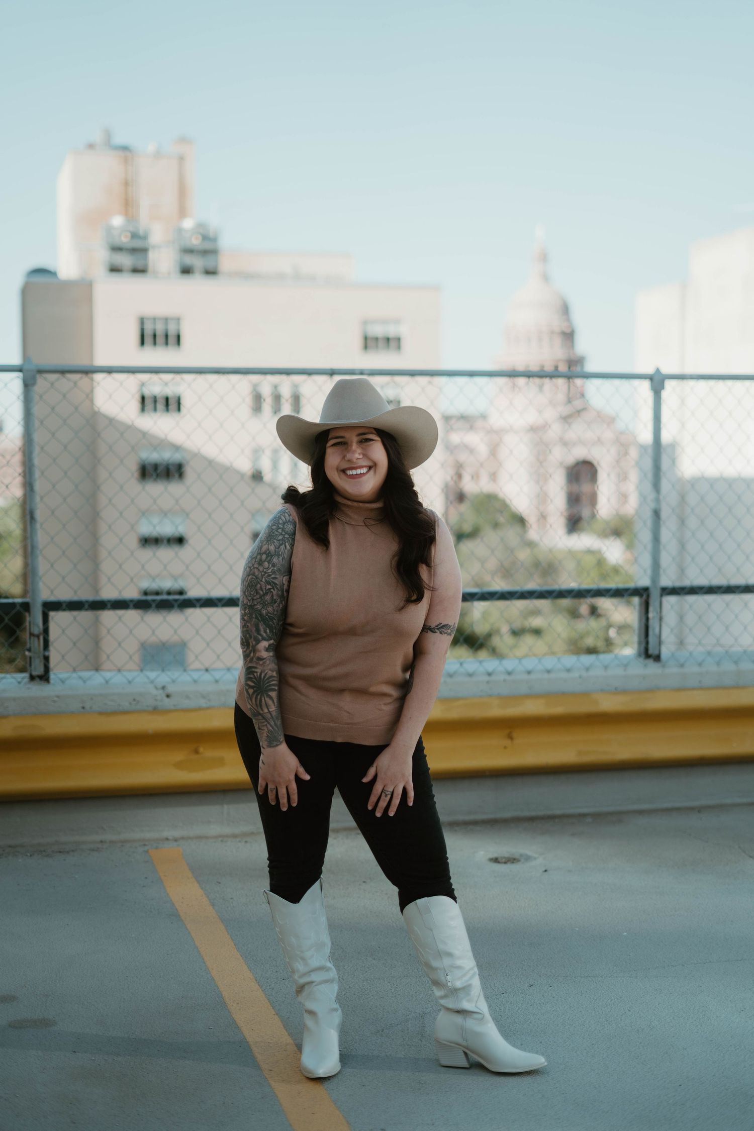 A person in a tan cowboy hat and boots poses on a rooftop parking deck with city buildings in the background.