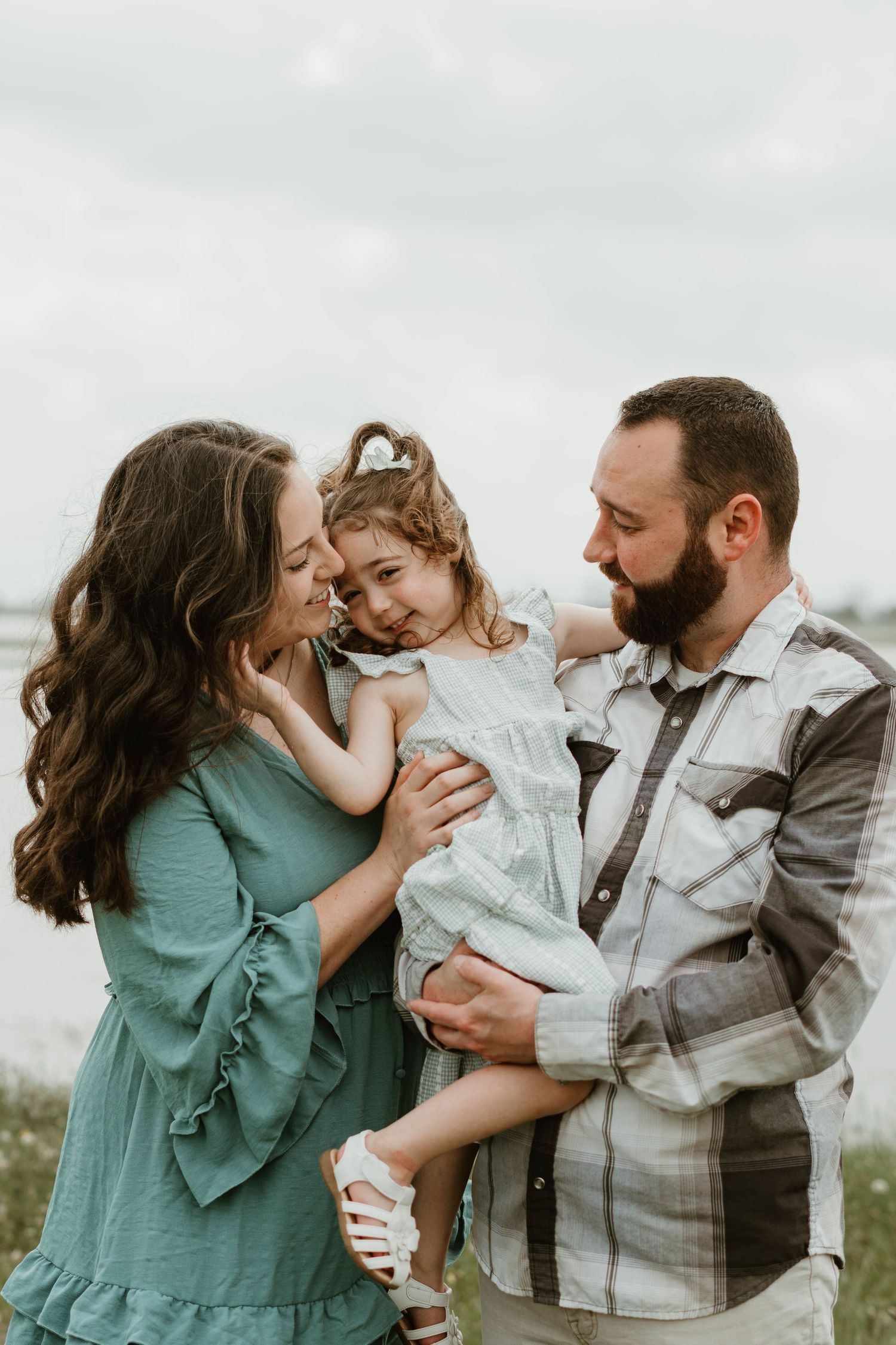 A happy family shares a tender moment together outdoors, with the child wearing a white dress and parents in casual attire.