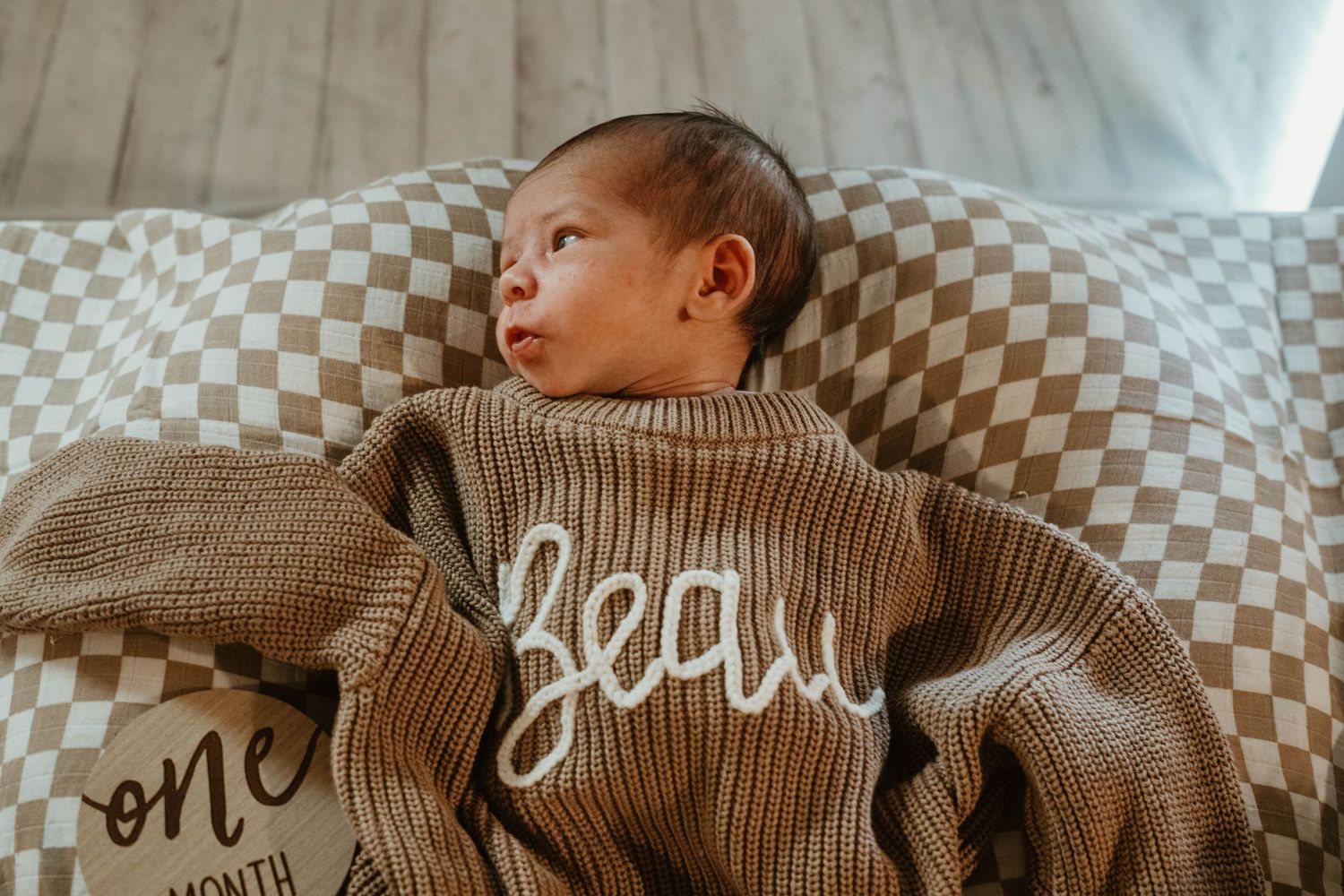 Baby wearing brown knit sweater lying on checkered grey and white blanket in soft natural light.
