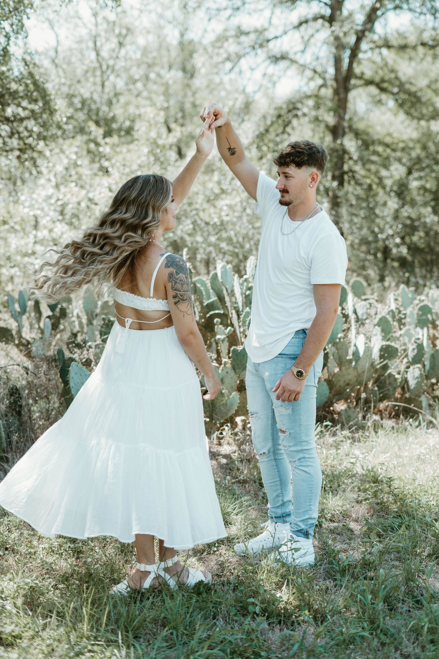 A romantic outdoor photoshoot with a couple in white clothing dancing among cactus plants in a dreamy sunlit setting.
