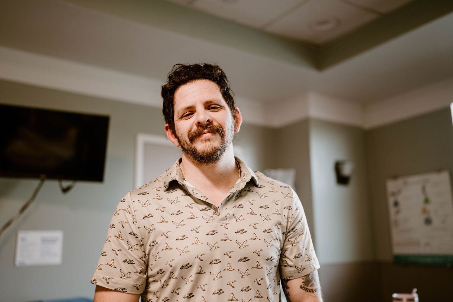 A person in a floral print shirt smiles while standing in an office environment.