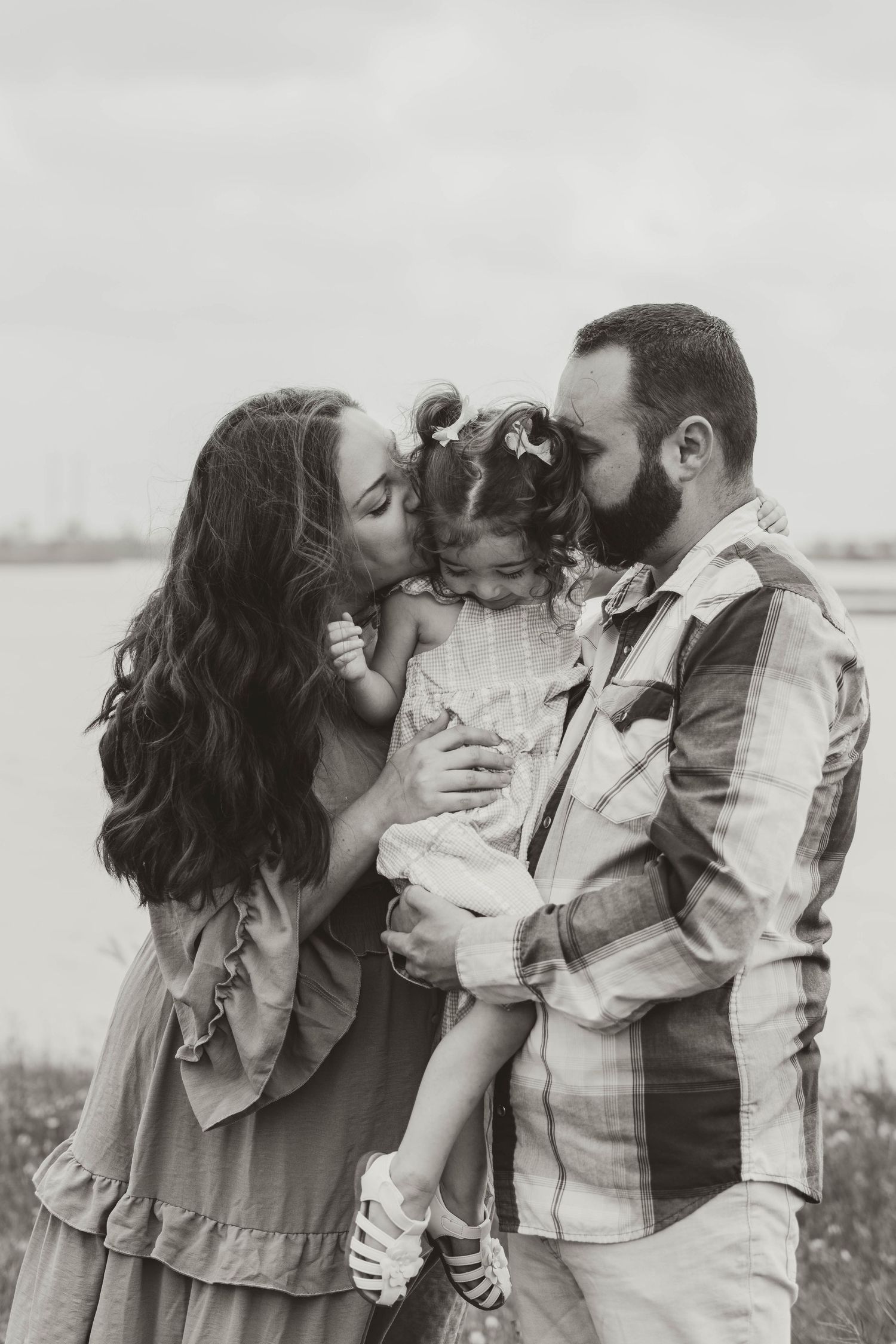 A tender black and white family moment outdoors as parents share kisses with their young child.