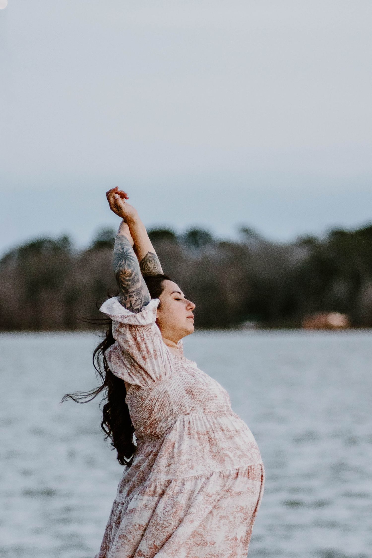 A person in a flowing pink floral dress twirls joyfully by a lake on an overcast day.