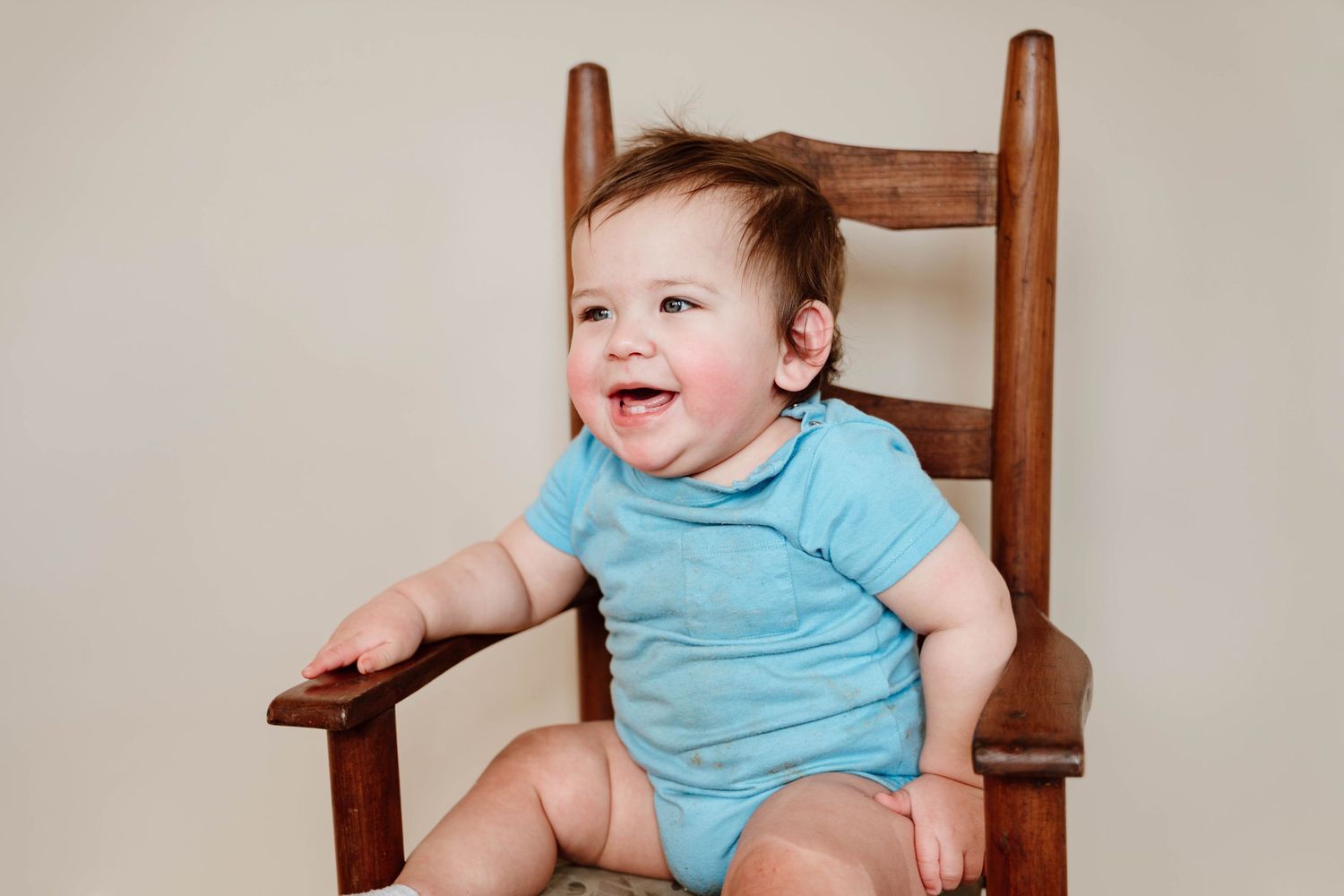 Happy baby in blue onesie sitting in wooden rocking chair, smiling and laughing against neutral background.
