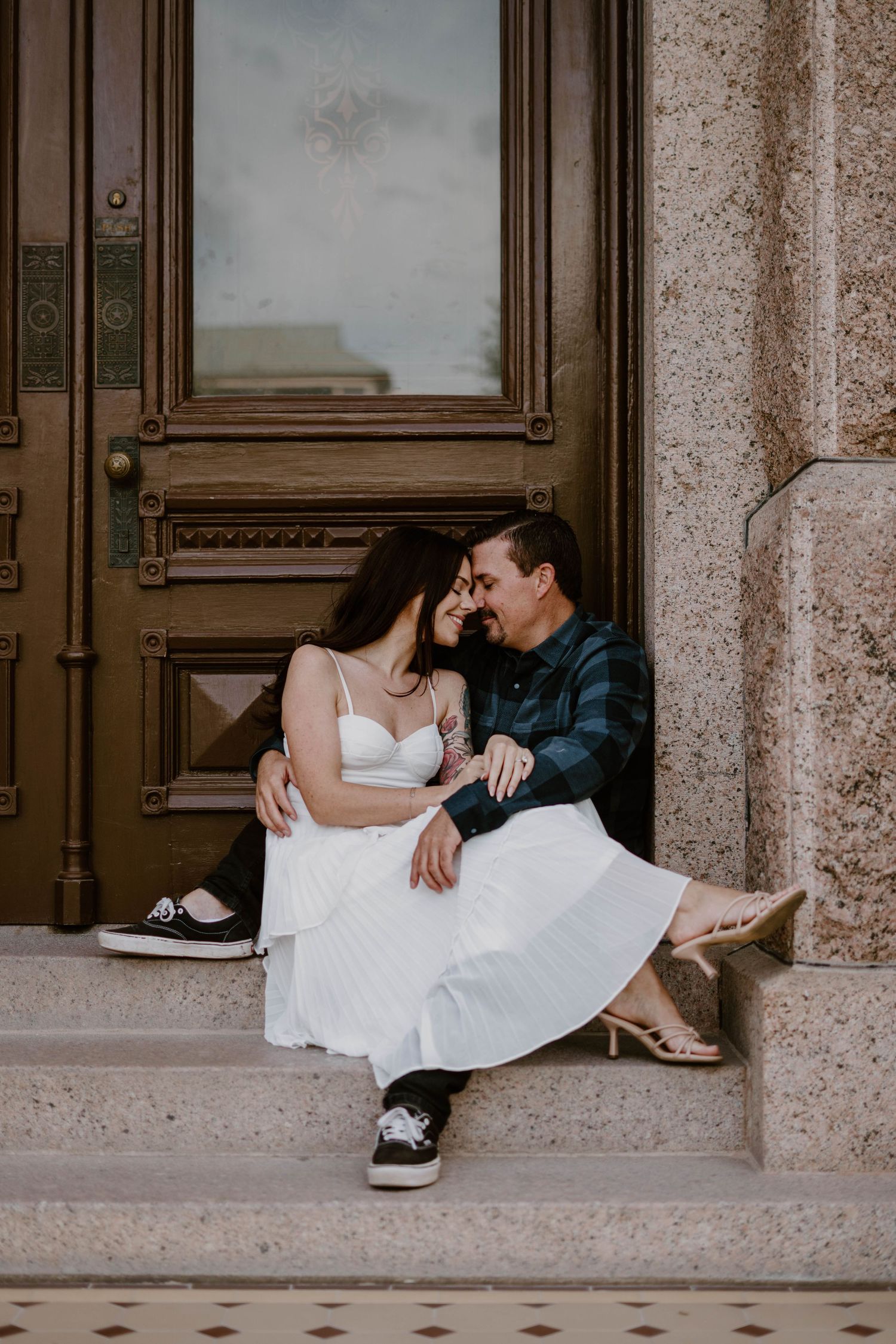 A romantic couple sits intimately on stone steps in front of a vintage wooden door, dressed in casual and formal attire.