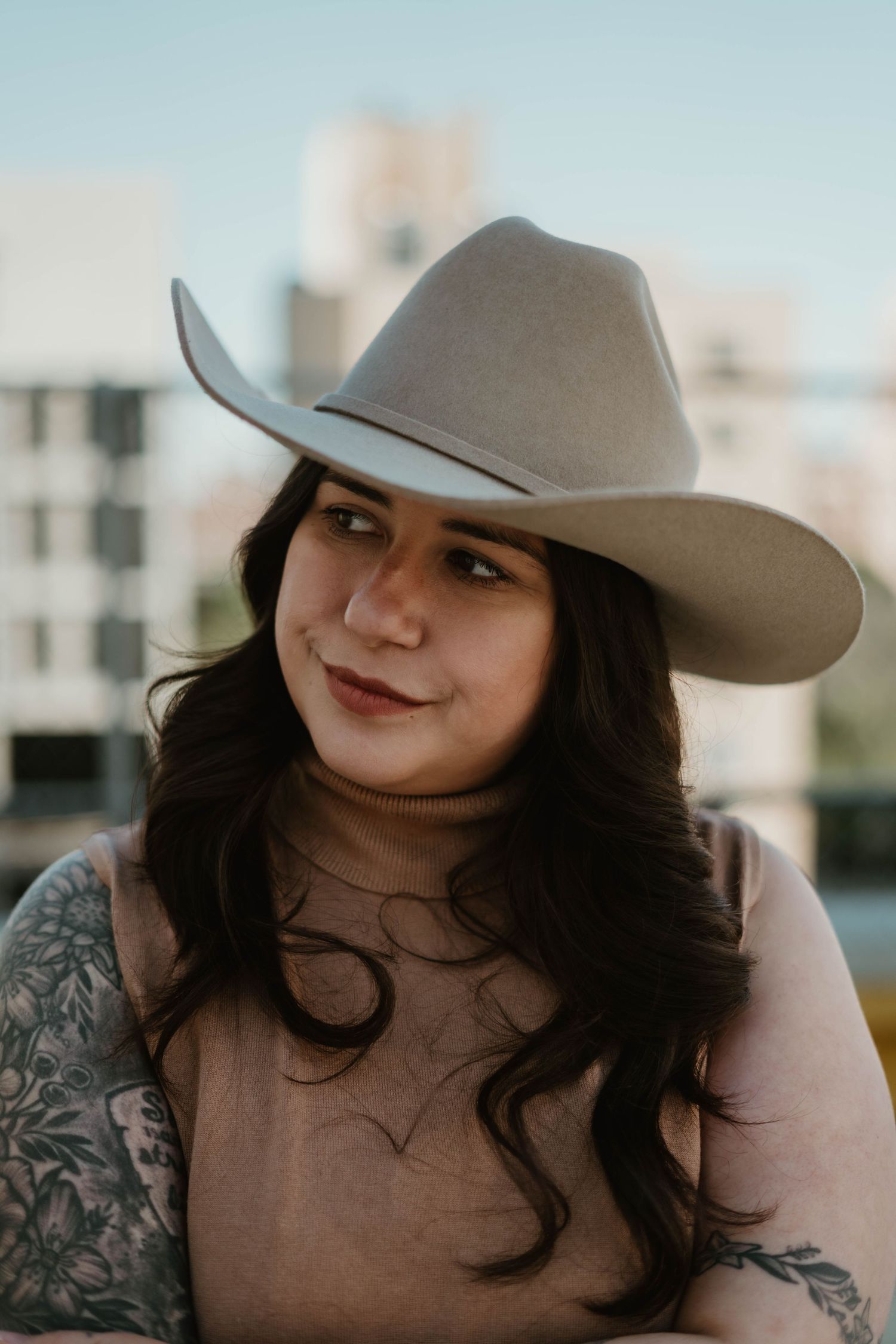 A person wearing a light gray cowboy hat and long dark hair smiles while showing visible arm tattoos.