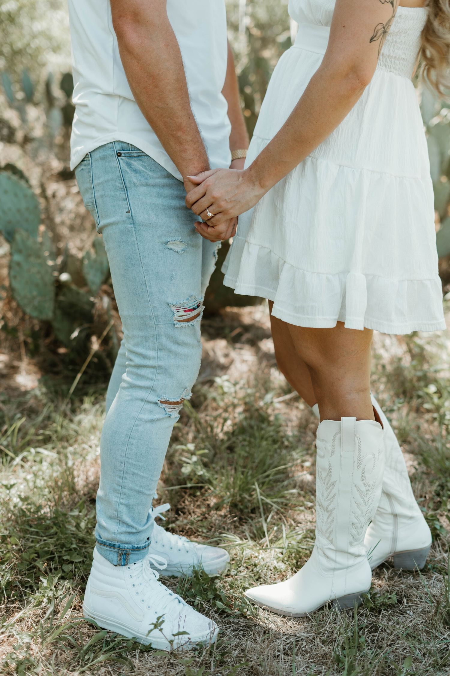 Couple in white outfits holding hands outdoors, with person in dress wearing white cowboy boots and other in ripped jeans.