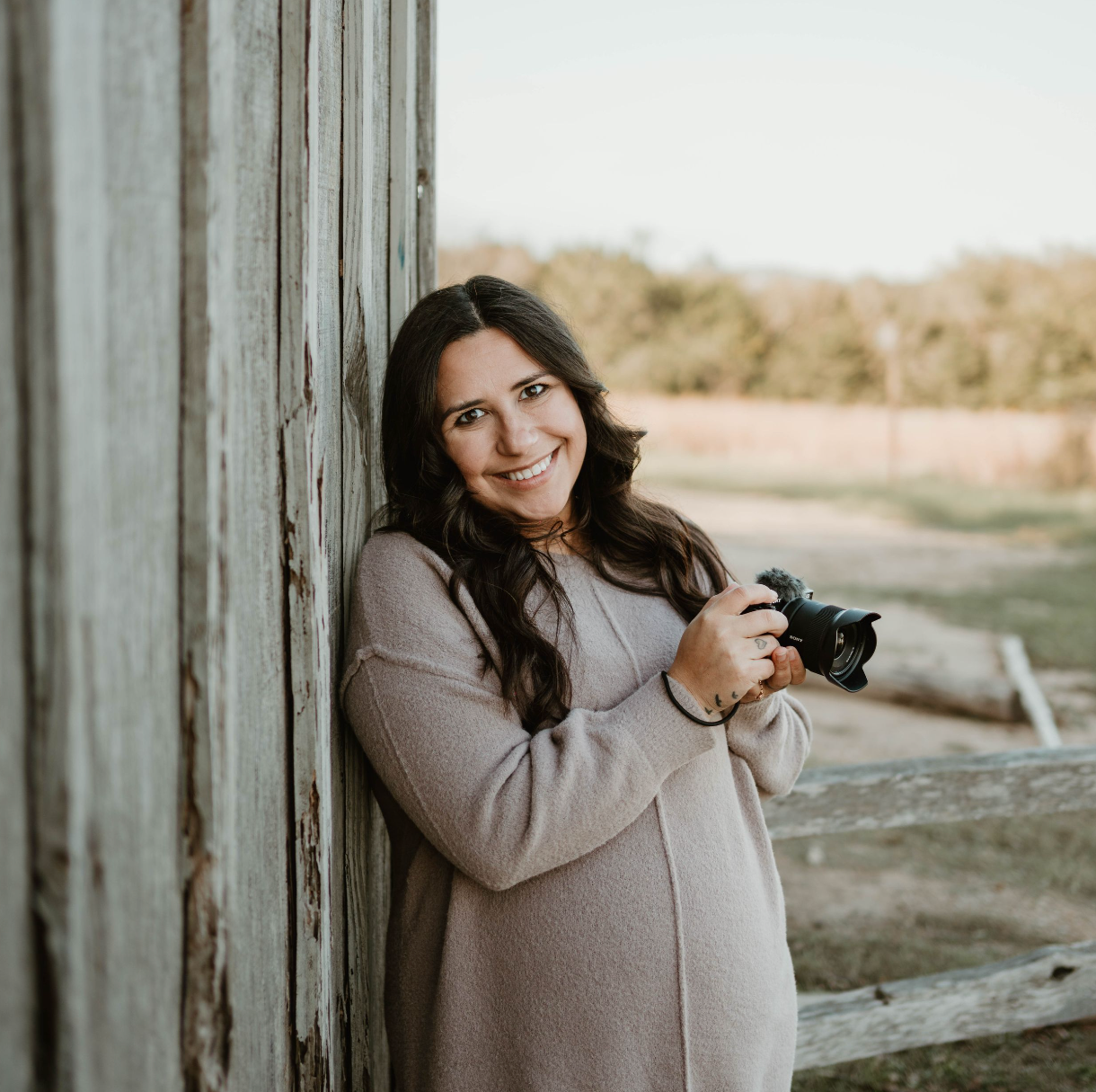 A photographer in a beige sweater stands against a rustic wooden wall holding a camera in a golden-hour outdoor setting.
