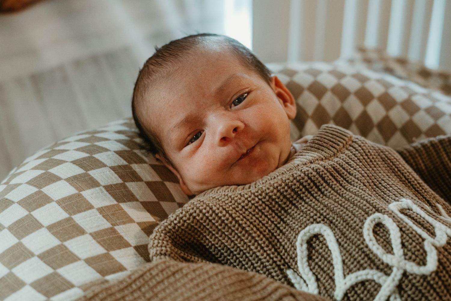 A small baby lying on patterned bedding while wearing a knitted sweater smiles at the camera.