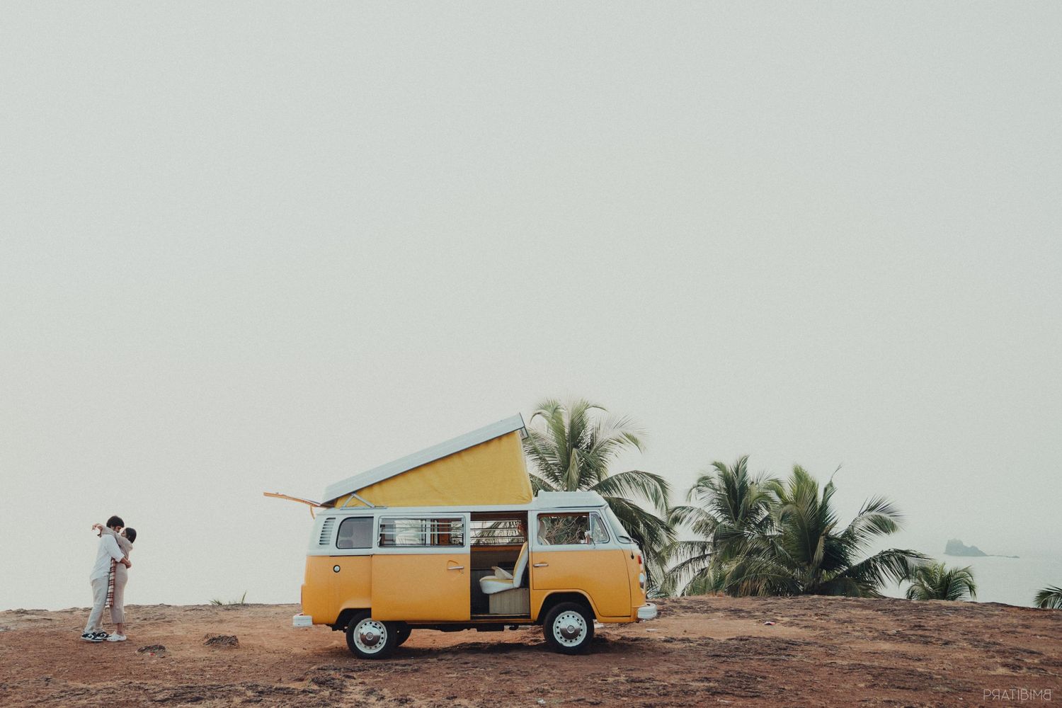 A yellow vintage VW van parked on a hilltop with palm trees in the background.