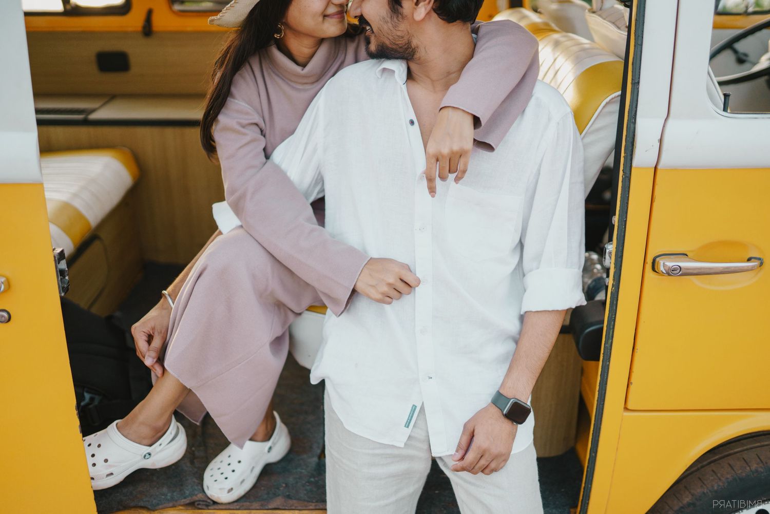 Couple in coordinated pink and white outfits share a tender moment by a yellow vintage bus.