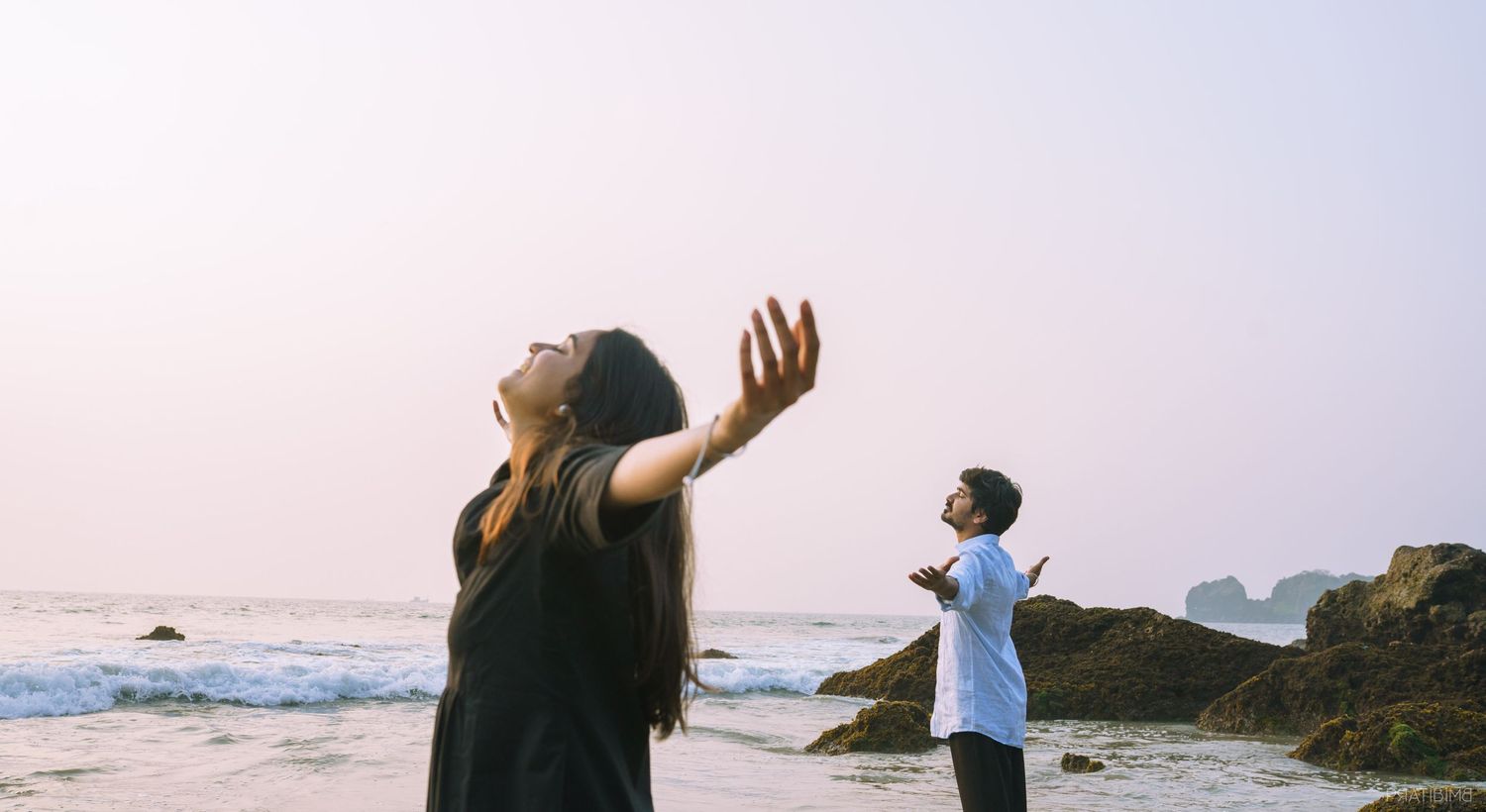 Two people strike joyful poses against an ocean backdrop during sunset.
