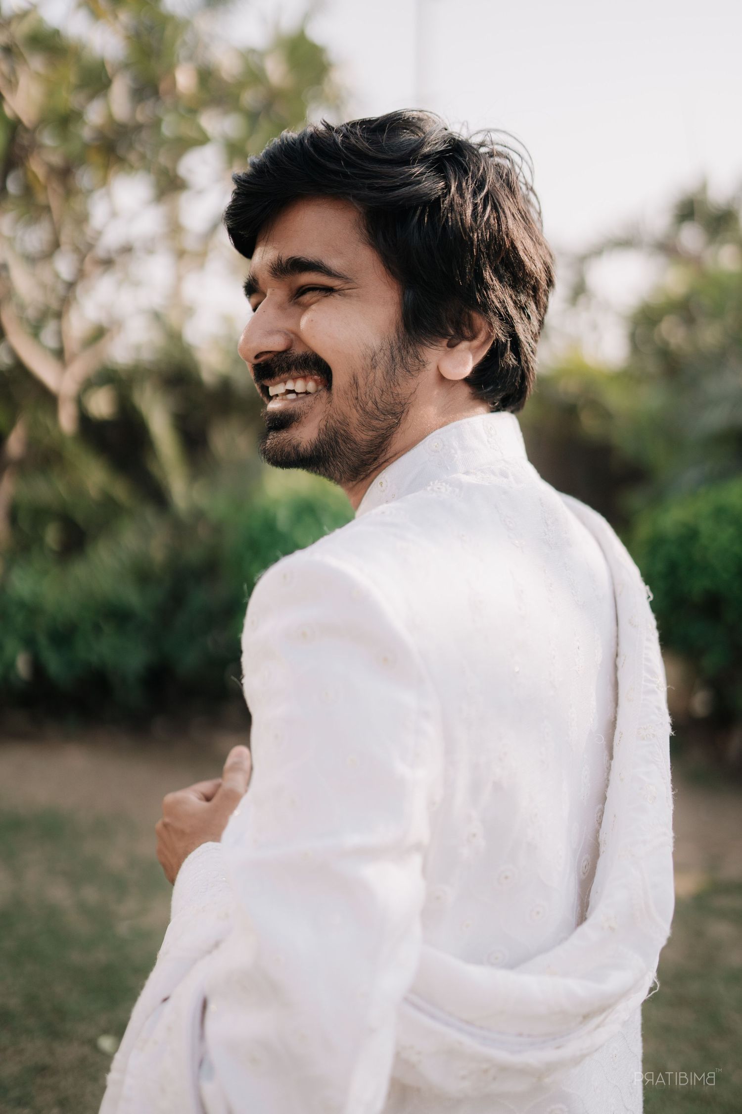 Person in white attire smiling against outdoor garden background.