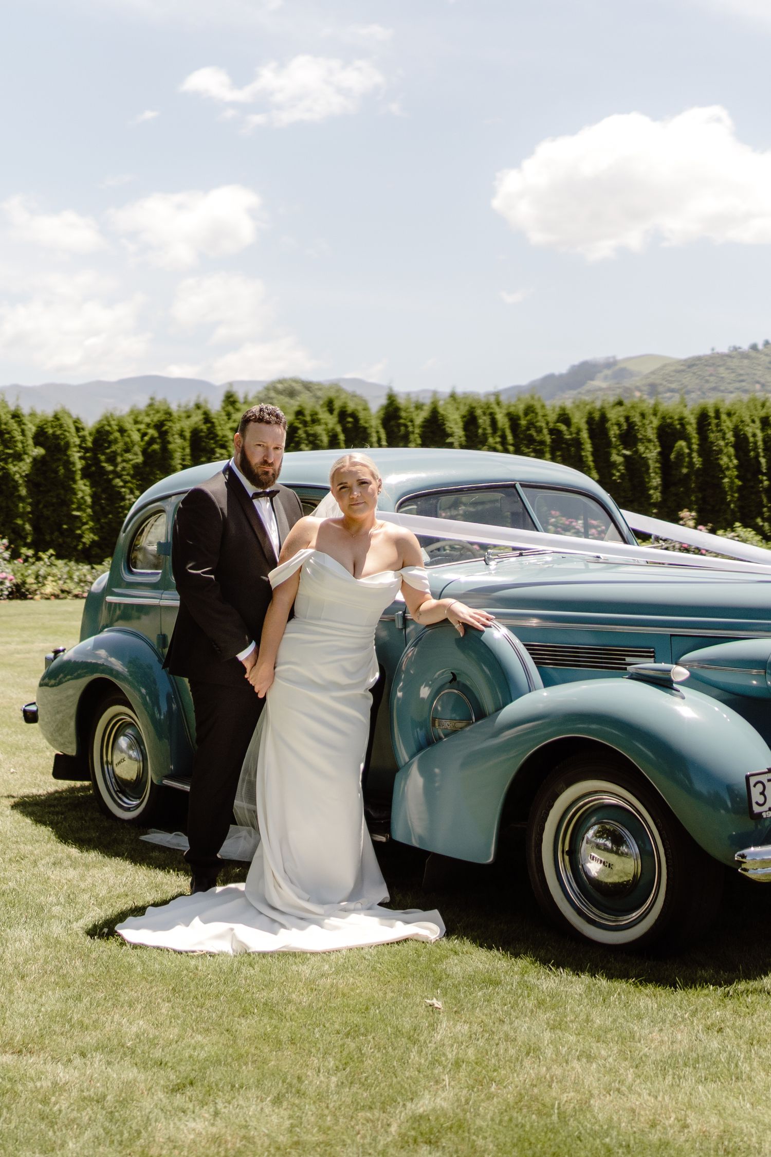 An editorial wedding photo of a bride and groom leaning on a blue vintage 1937 Buick car with a stunning green backdrop with rolling hills