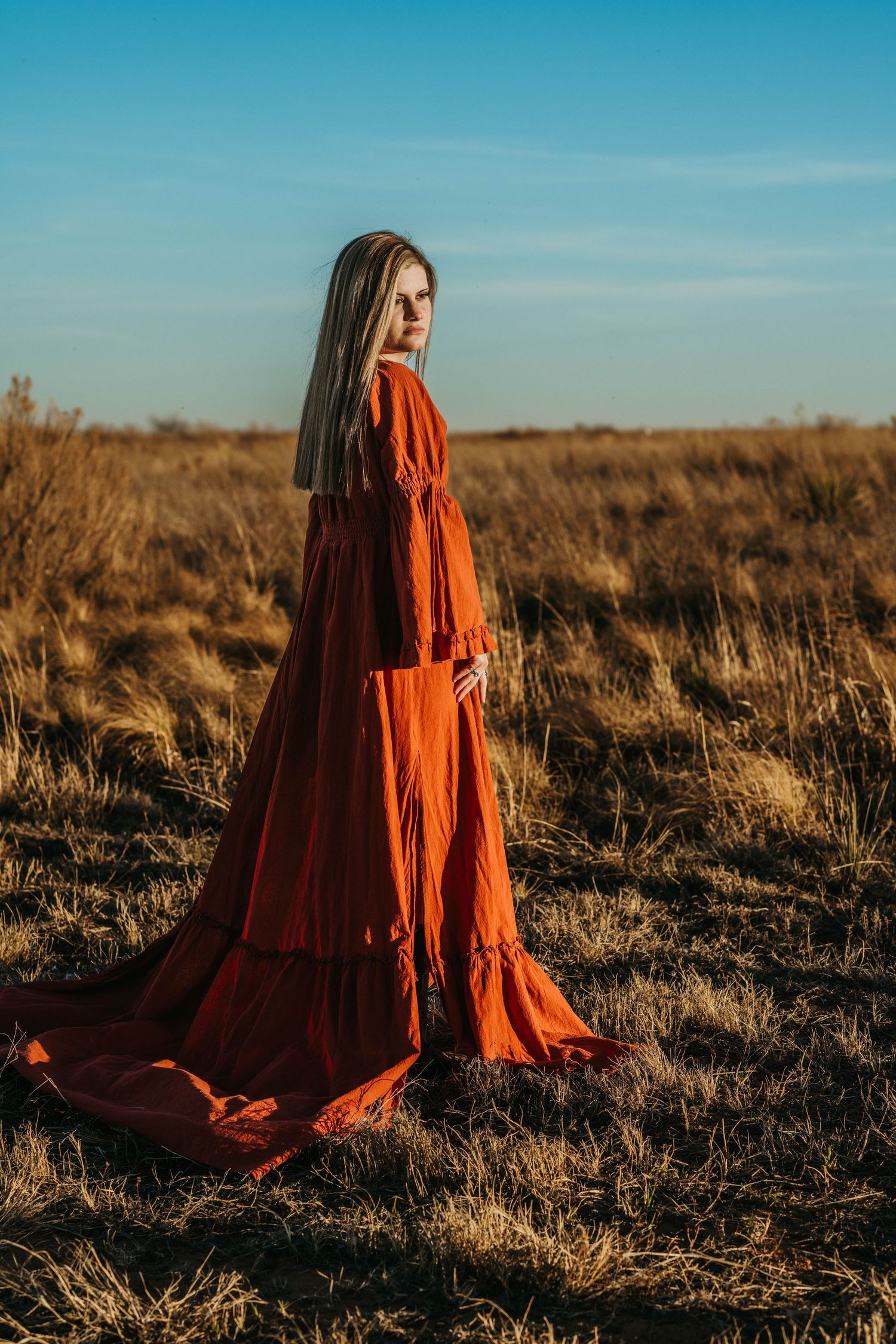 A flowing red dress billows dramatically against a golden field at sunset with blue sky overhead.
