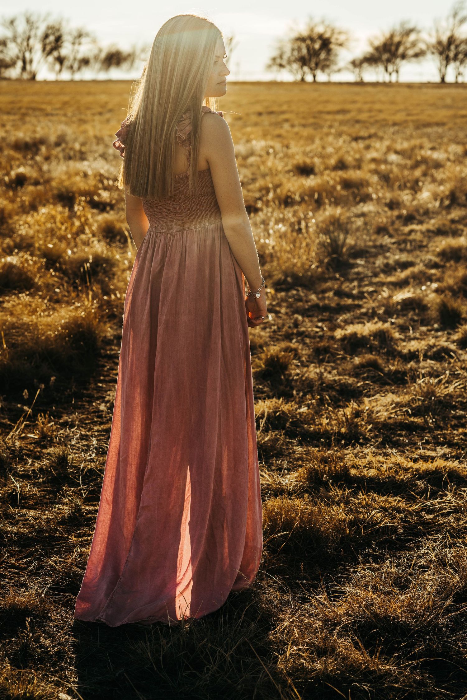 A figure in a flowing pink dress stands in a golden field at sunset, creating a dreamy silhouette against the landscape.