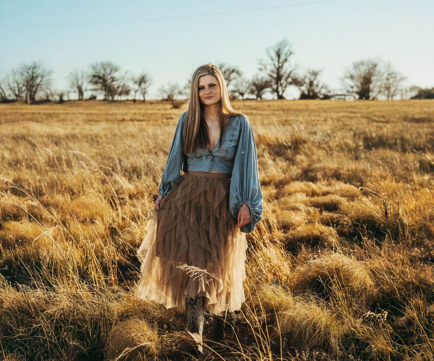A person in a blue top and flowing skirt stands in a golden autumn field at sunset with bare trees in the background.