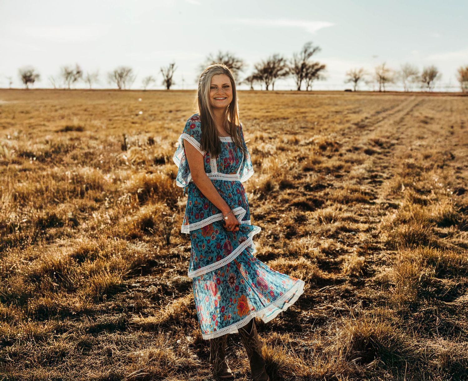A woman in a flowy blue dress twirls gracefully in a golden field at sunset.