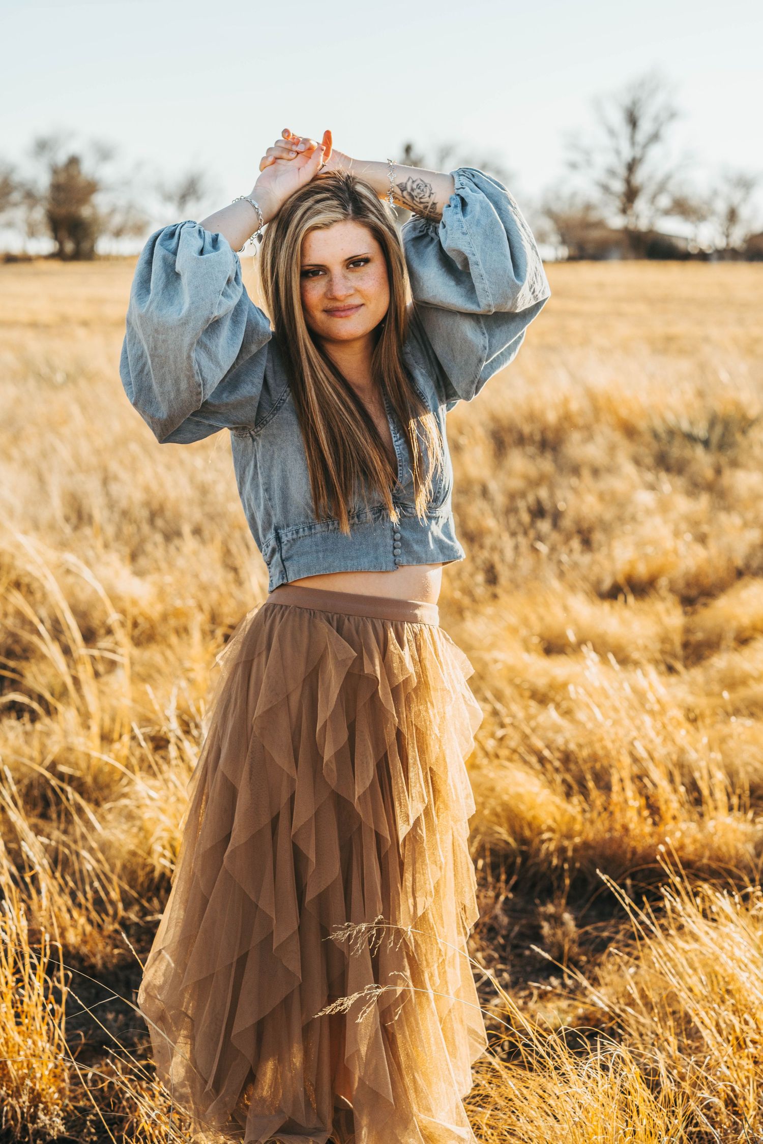 A person in a denim jacket and tulle skirt poses playfully in a golden field at sunset.