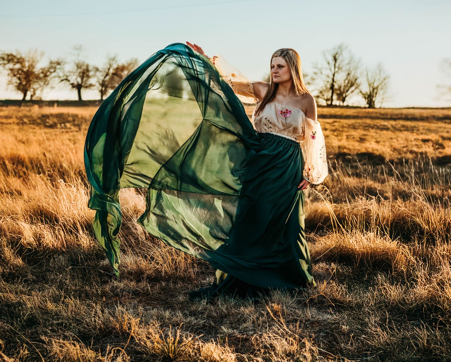A flowing emerald green dress billows dramatically in the golden light of a sunset field.