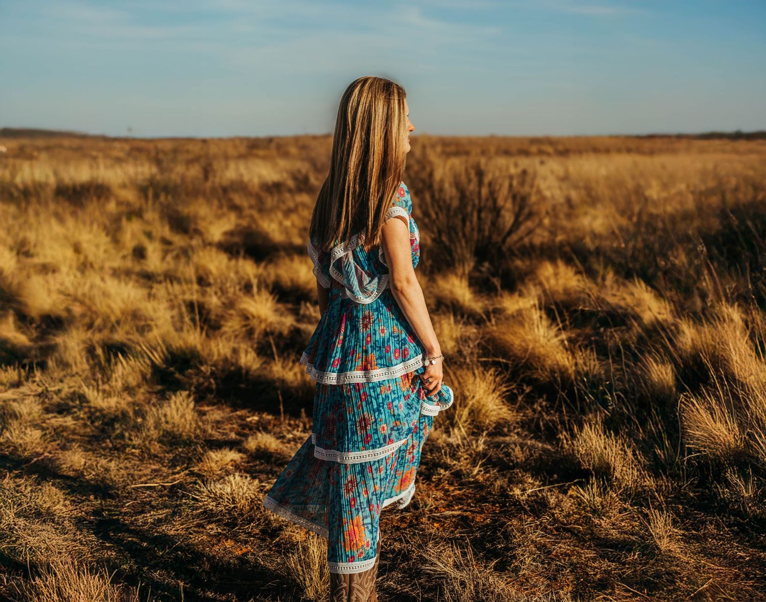 A figure in a plaid dress stands in a golden field at sunset, photographed from behind.