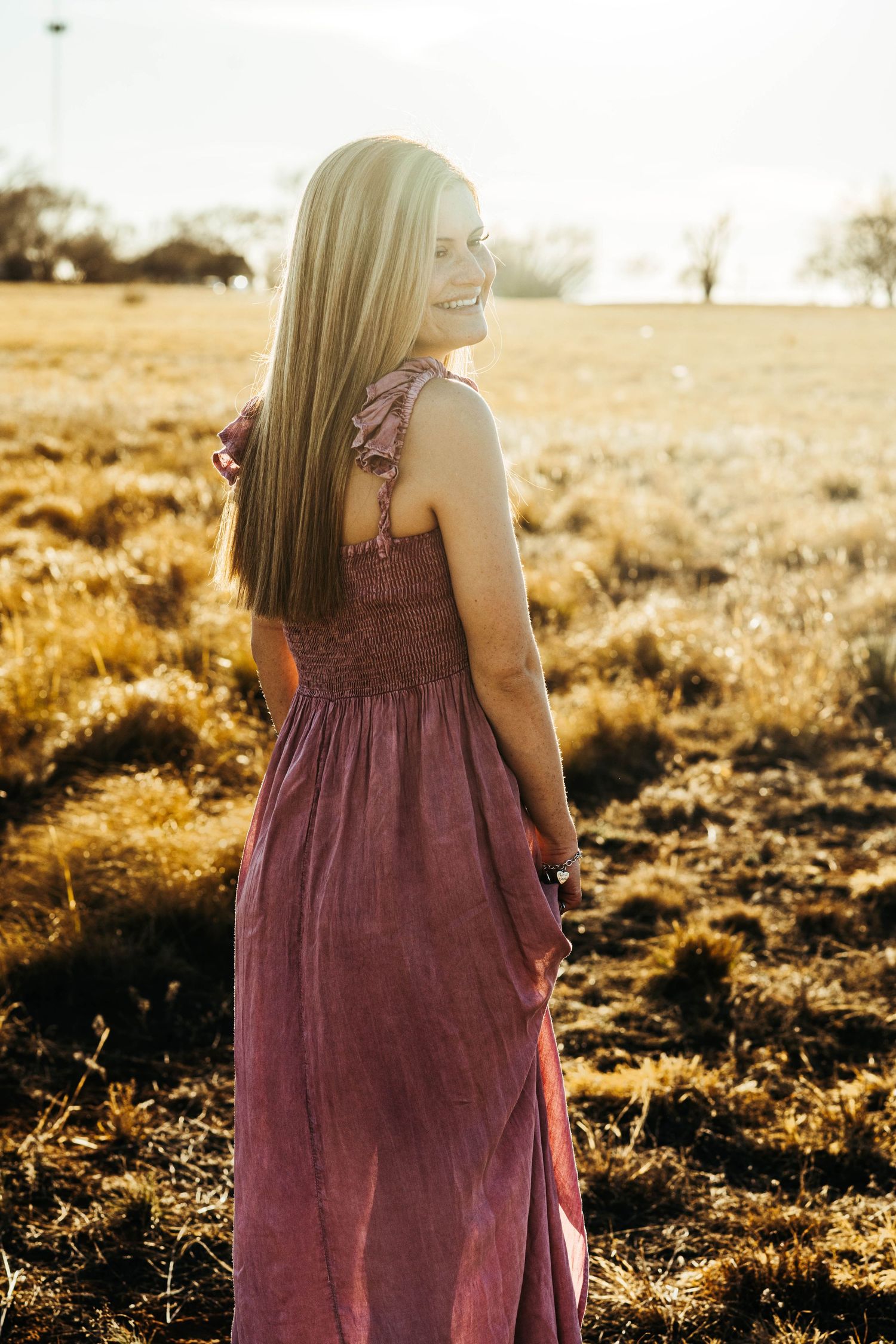 A person in a mauve dress stands in a golden sunlit field during sunset, photographed from behind.