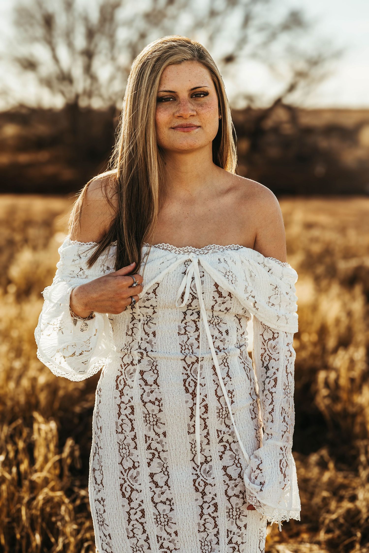 A person wearing a white lace off-shoulder dress poses in a golden field at sunset.