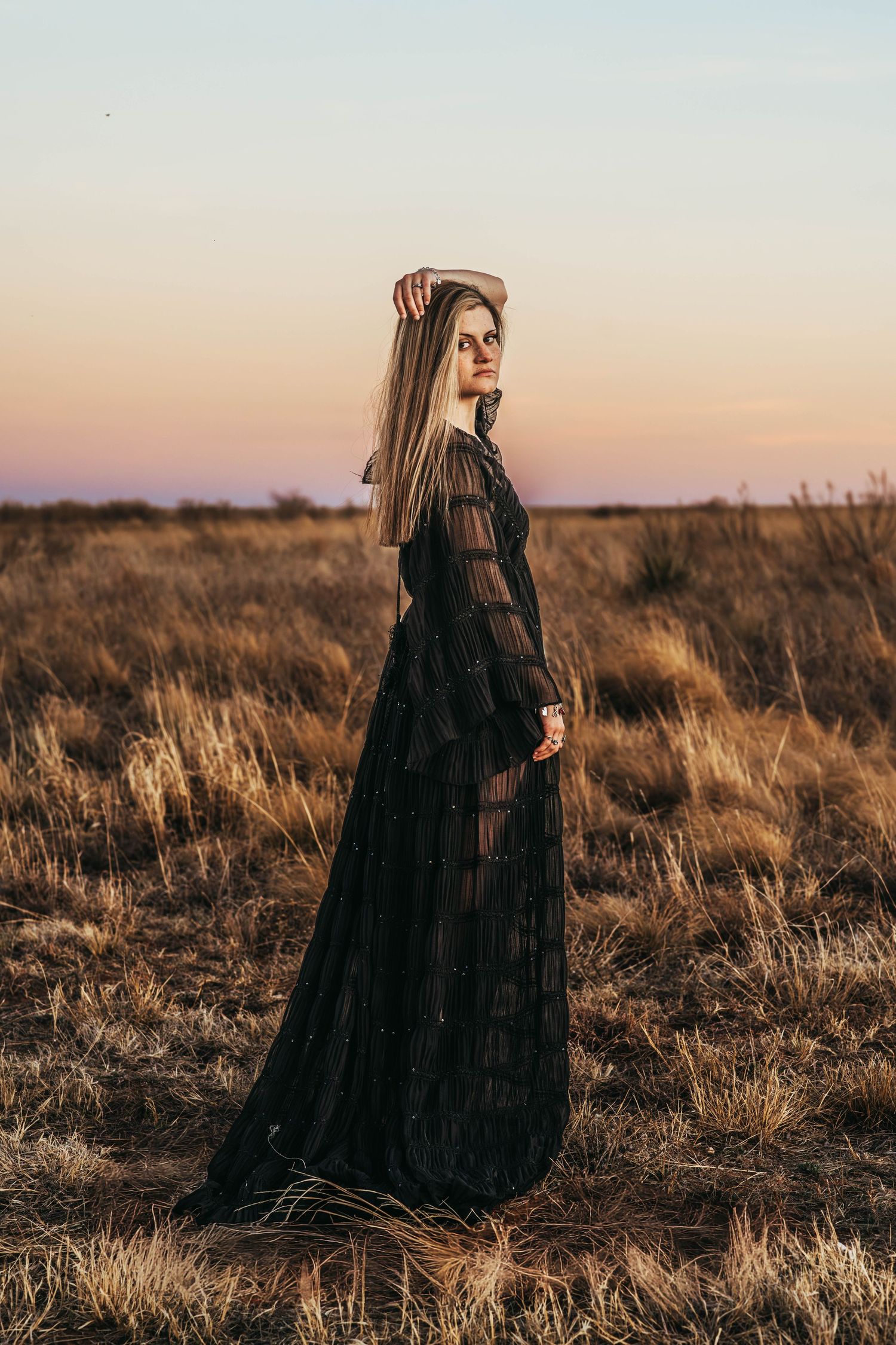 A figure in a long black Victorian-style dress stands in a golden field at sunset against a pastel sky.