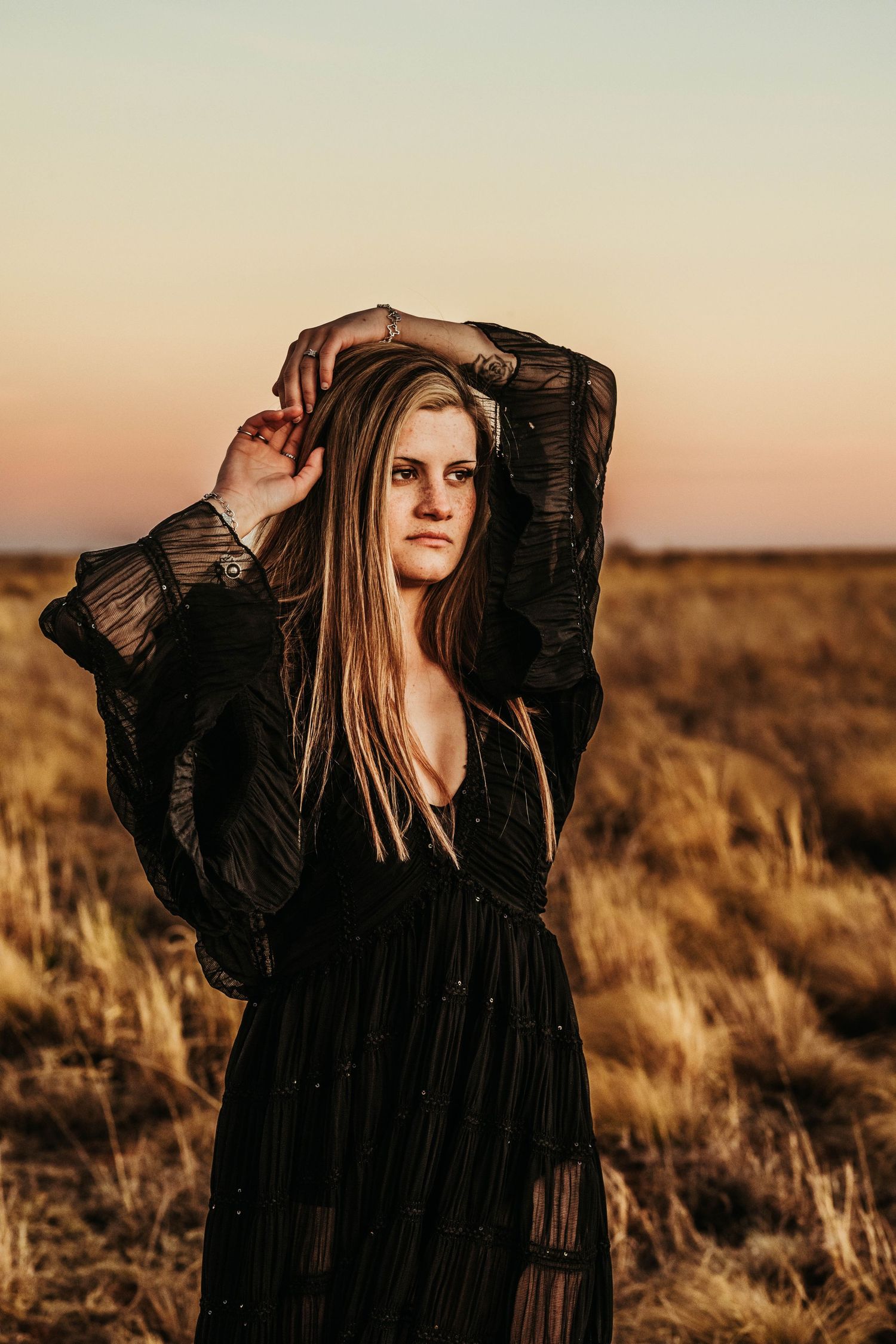 A figure in a flowing black dress poses dramatically against a golden sunset in tall grass prairie landscape.