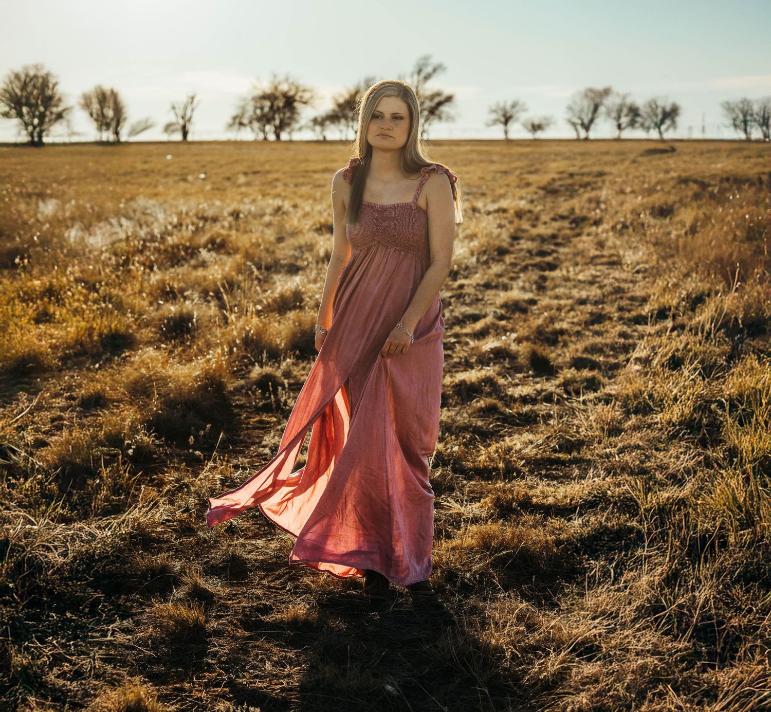 A figure in a flowing pink dress stands in a golden field at sunset with silhouetted trees in the background.