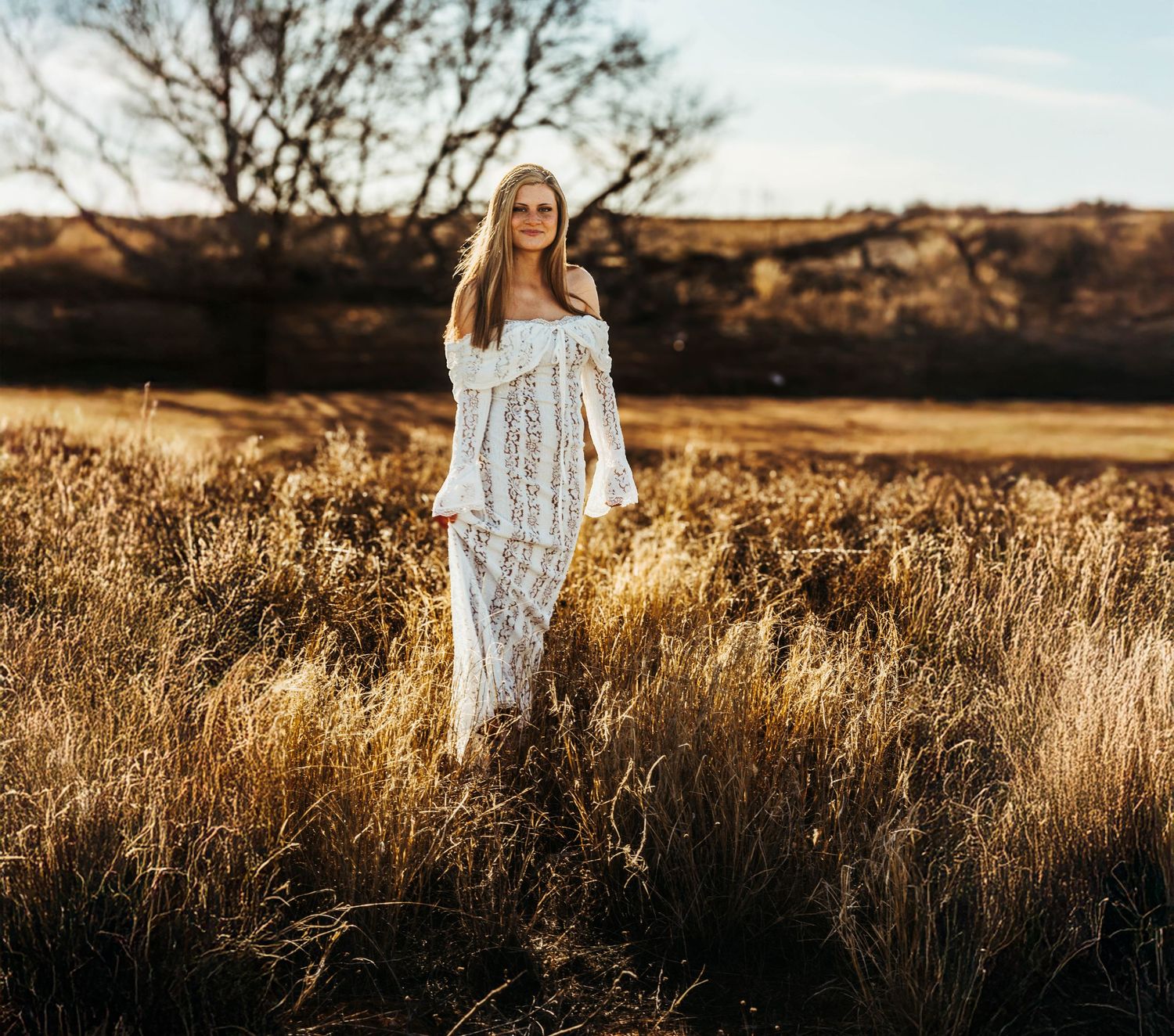 A person in a flowing white dress stands among golden grass in a rustic field at sunset.
