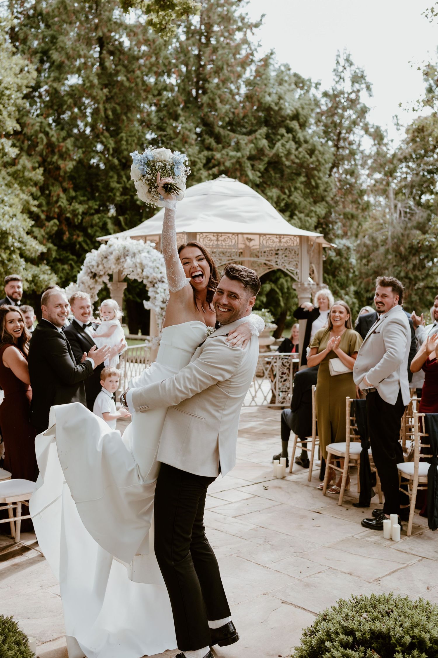 Newlyweds celebrate joyfully during their outdoor garden wedding ceremony as guests applaud in the background.