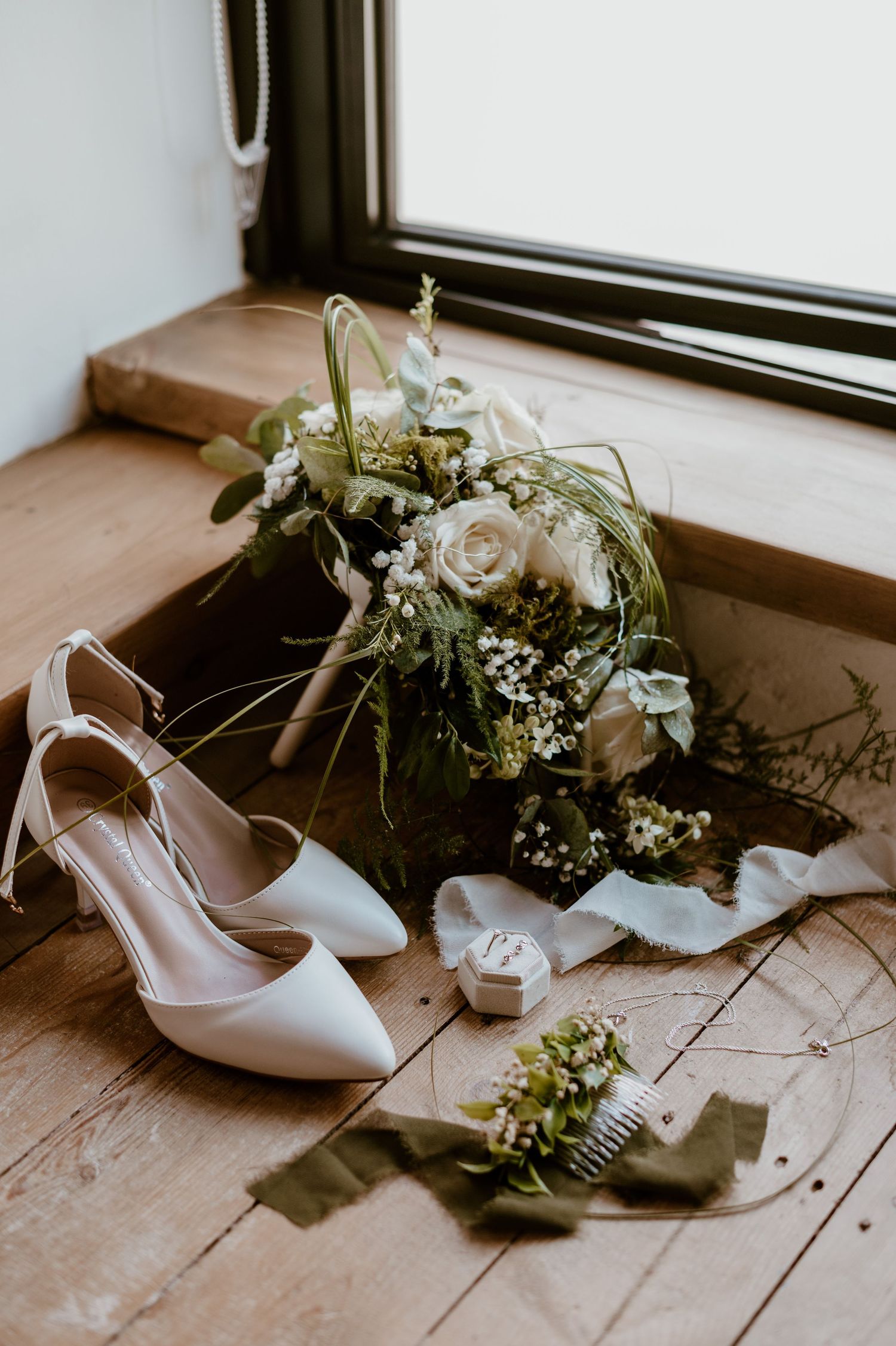 White wedding shoes and bridal bouquet with roses and greenery arranged on wooden windowsill in natural light.