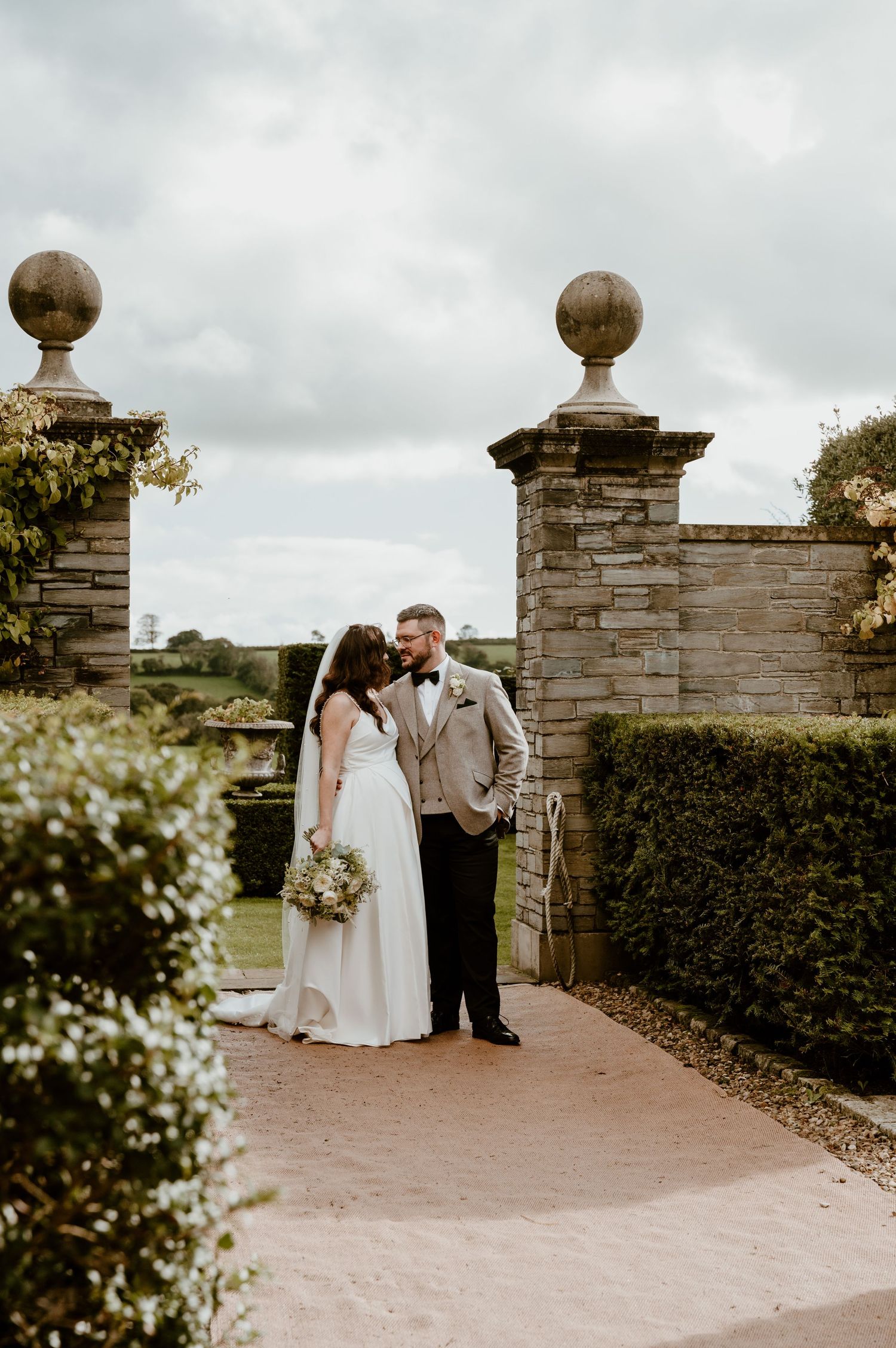 A couple in wedding attire shares intimate moments by a stone pillar entrance in a garden setting.