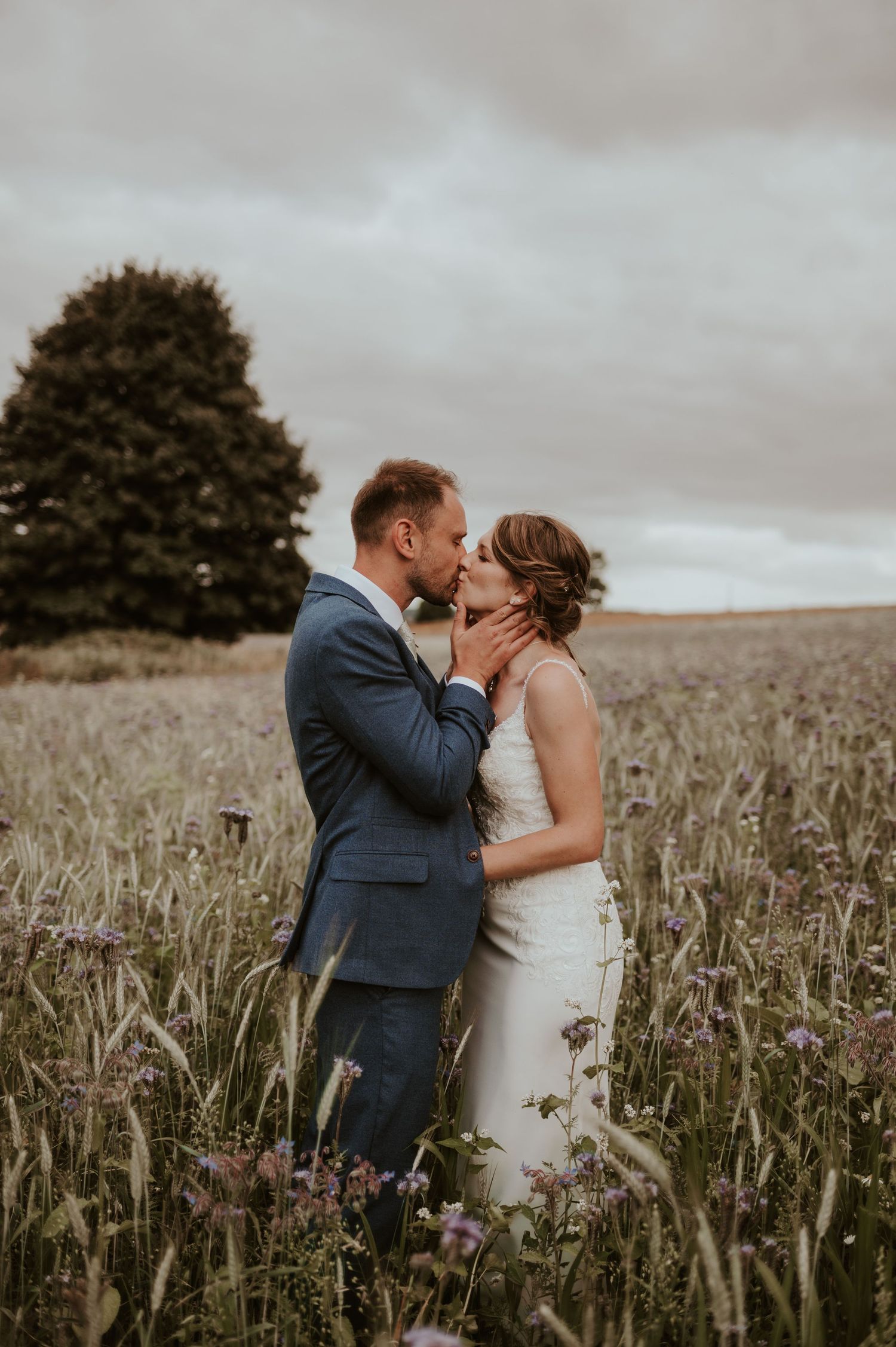 Newlywed couple shares an intimate moment in a wildflower field beneath moody clouds during sunset.