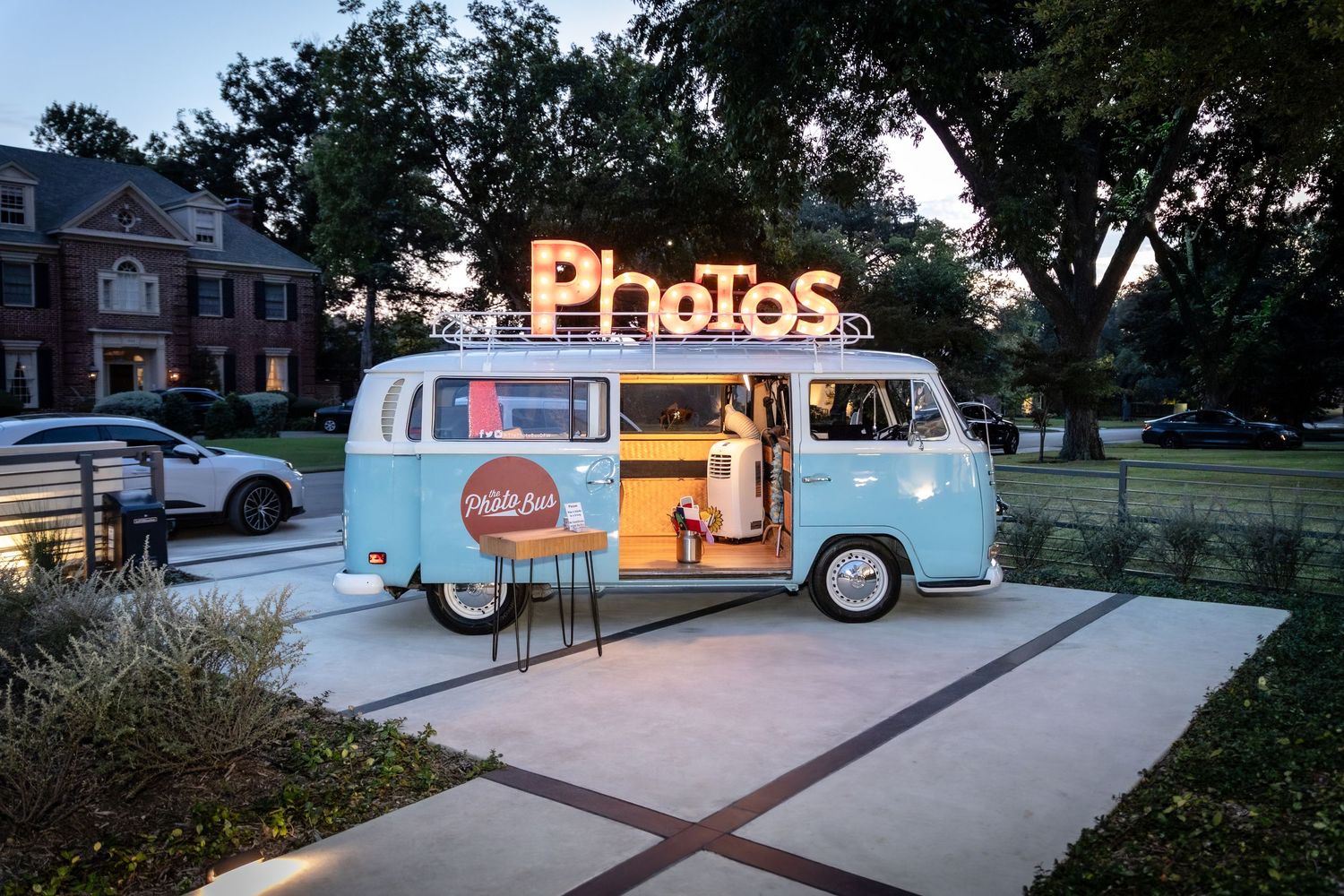 Vintage VW bus photo booth setup with illuminated signage at outdoor wedding venue at dusk.