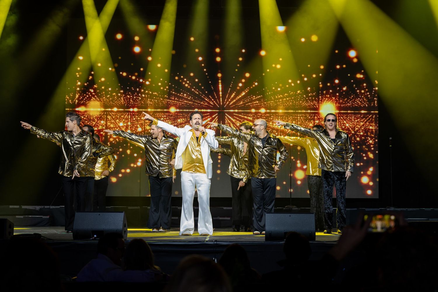 Concert performers on stage under dramatic yellow lighting with spotlights and pyrotechnic effects creating a spectacular atmosphere.