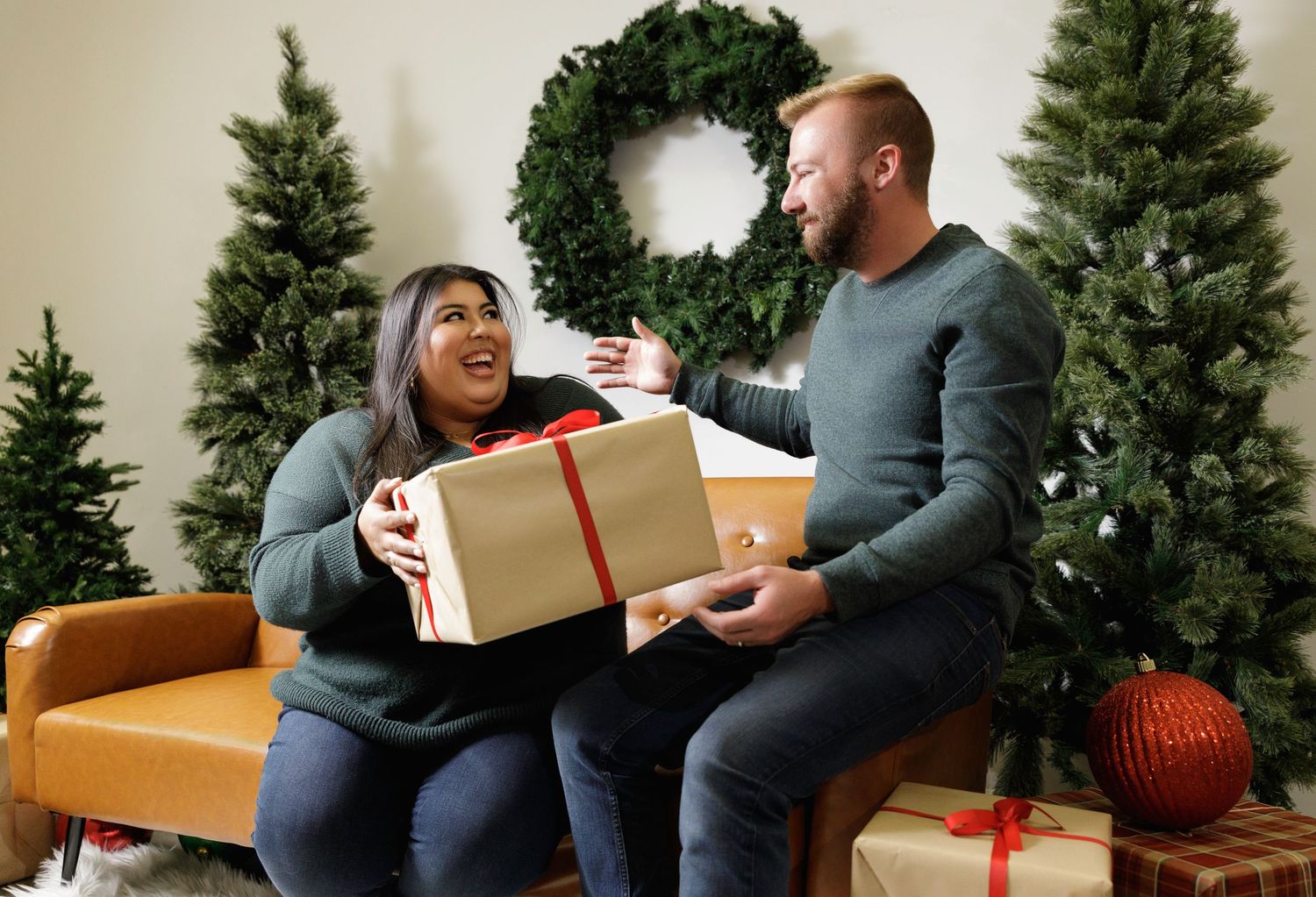 Couple opening Christmas presents together surrounded by decorated evergreen trees.