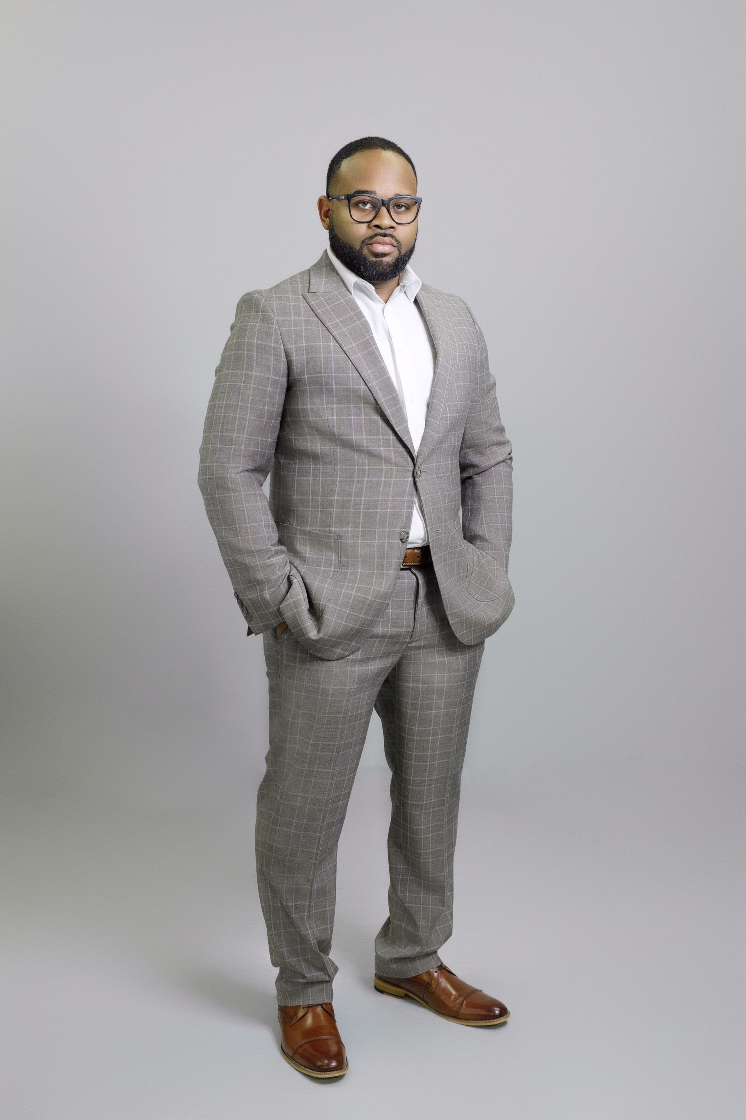 Professional wearing a well-tailored grey plaid suit with brown dress shoes poses against grey background.