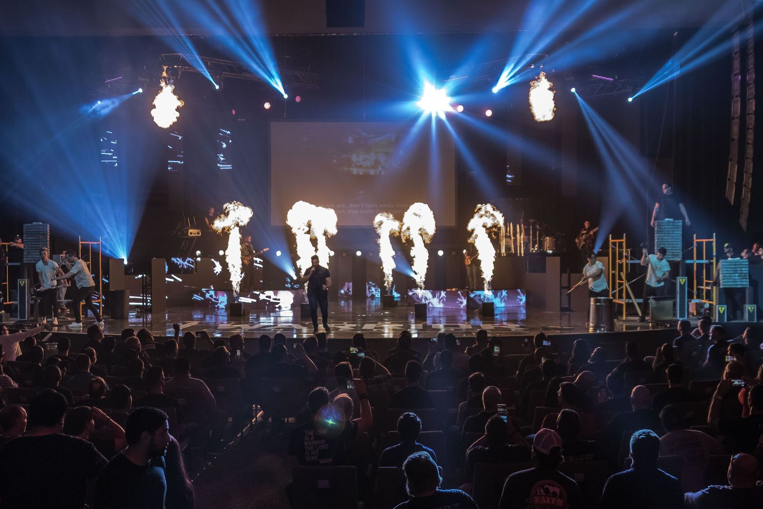 Concert stage illuminated with bright blue spotlights and fire effects during a live performance.