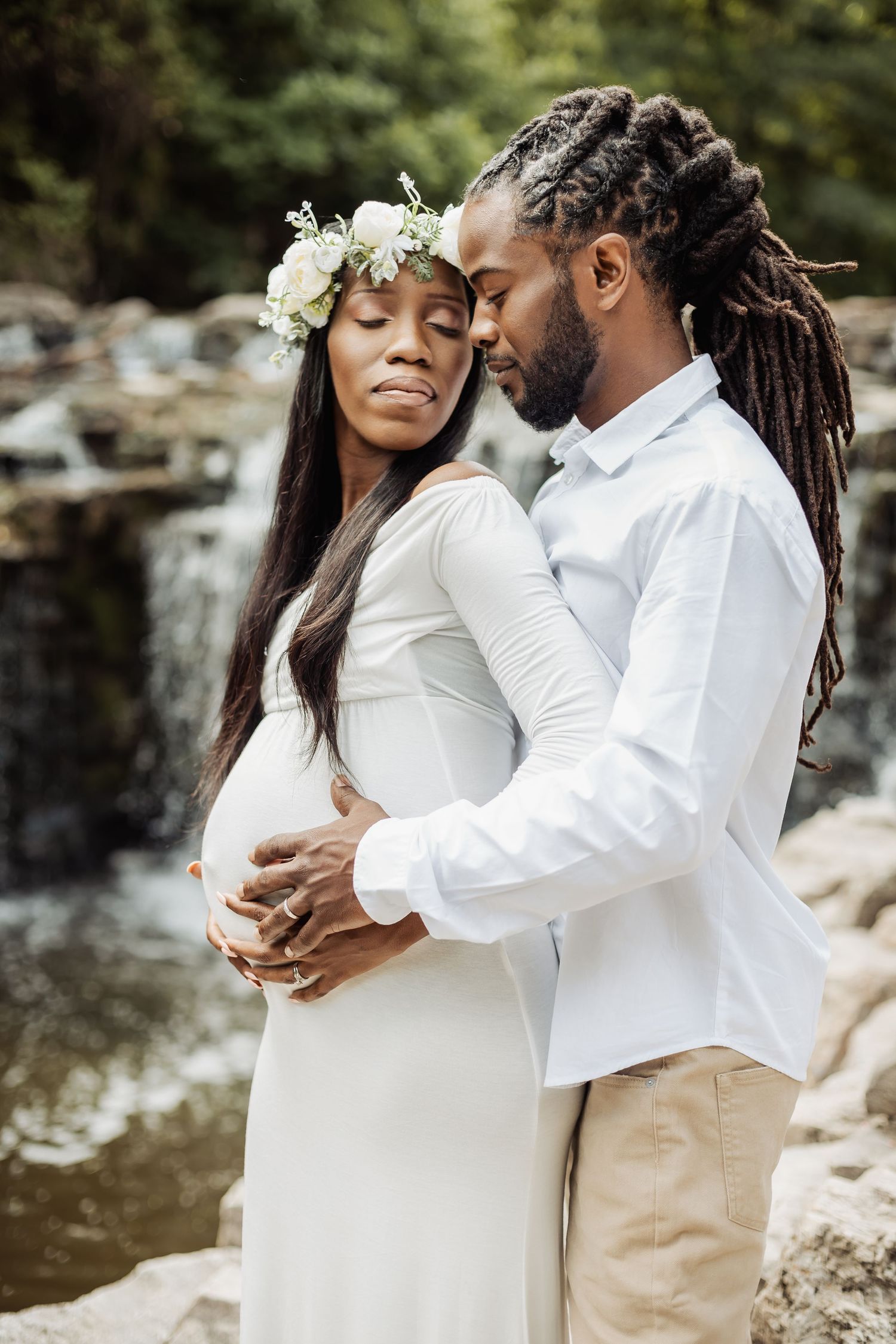 A romantic maternity photoshoot by a waterfall shows a couple in white and beige embracing tenderly outdoors.