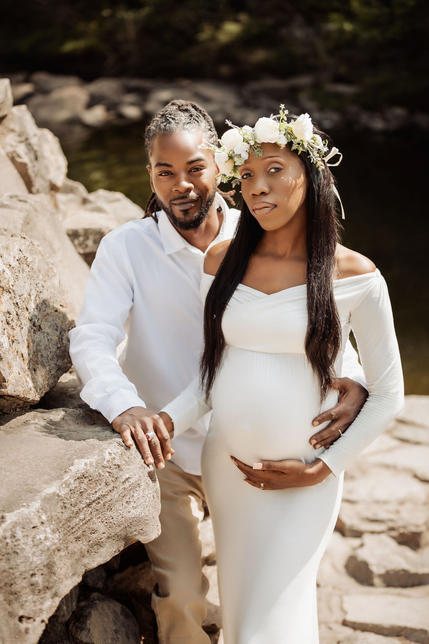 Couple in white outfits poses together for romantic maternity photos against a rocky outdoor backdrop.