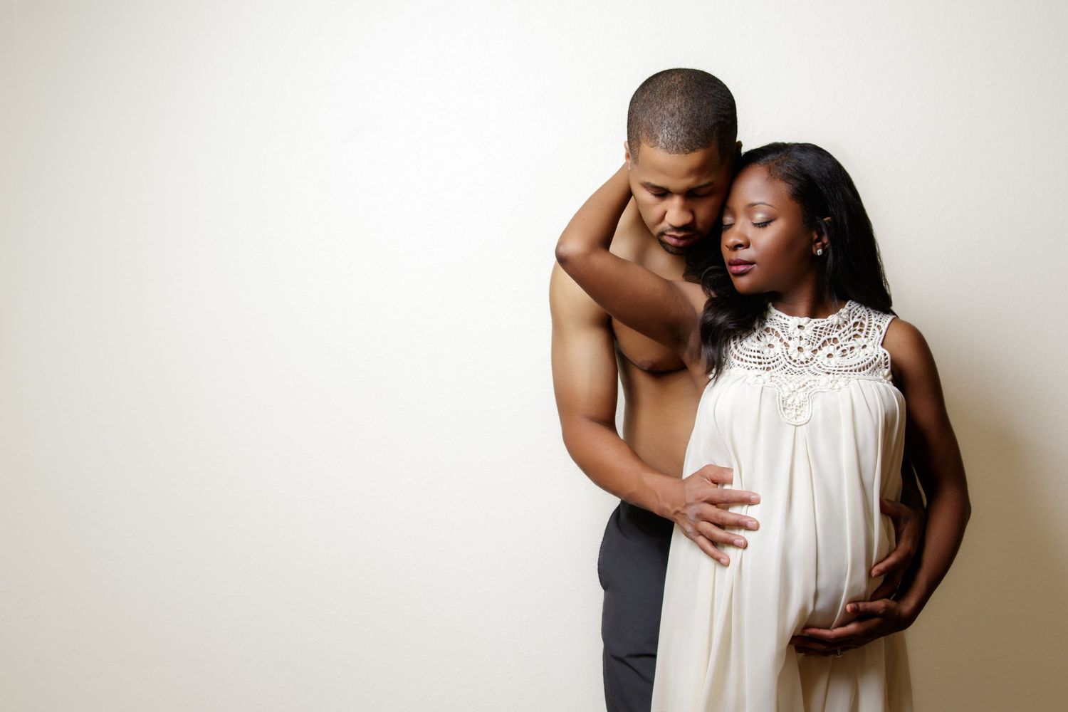 Artistic maternity photo showing couple in loving pose with white dress against light background.