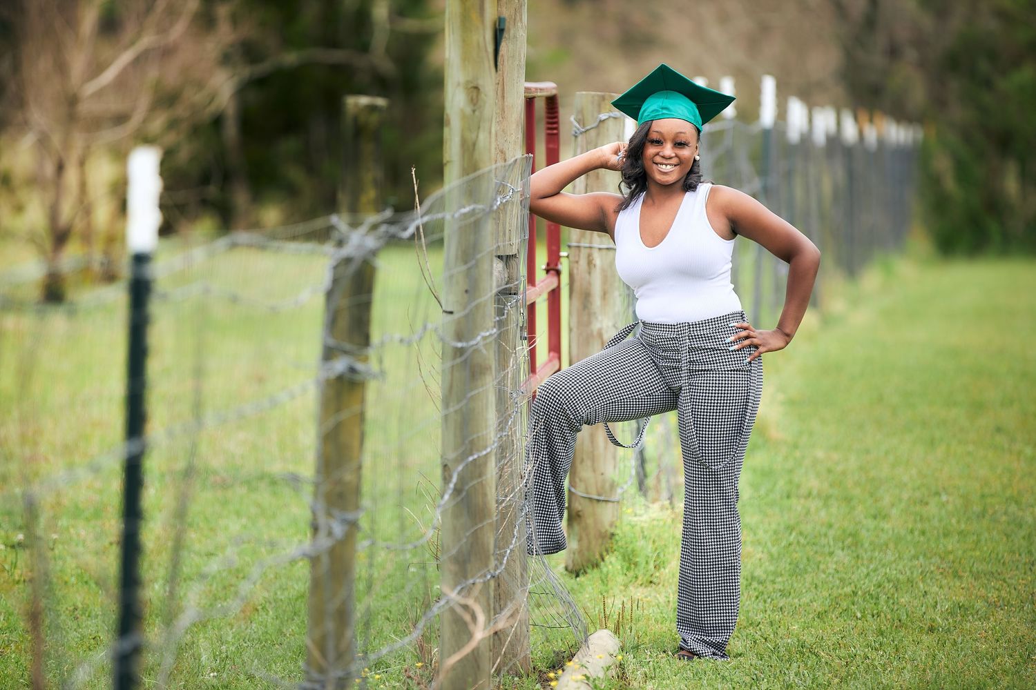 Person in casual attire poses playfully by red metal fence in rural setting.
